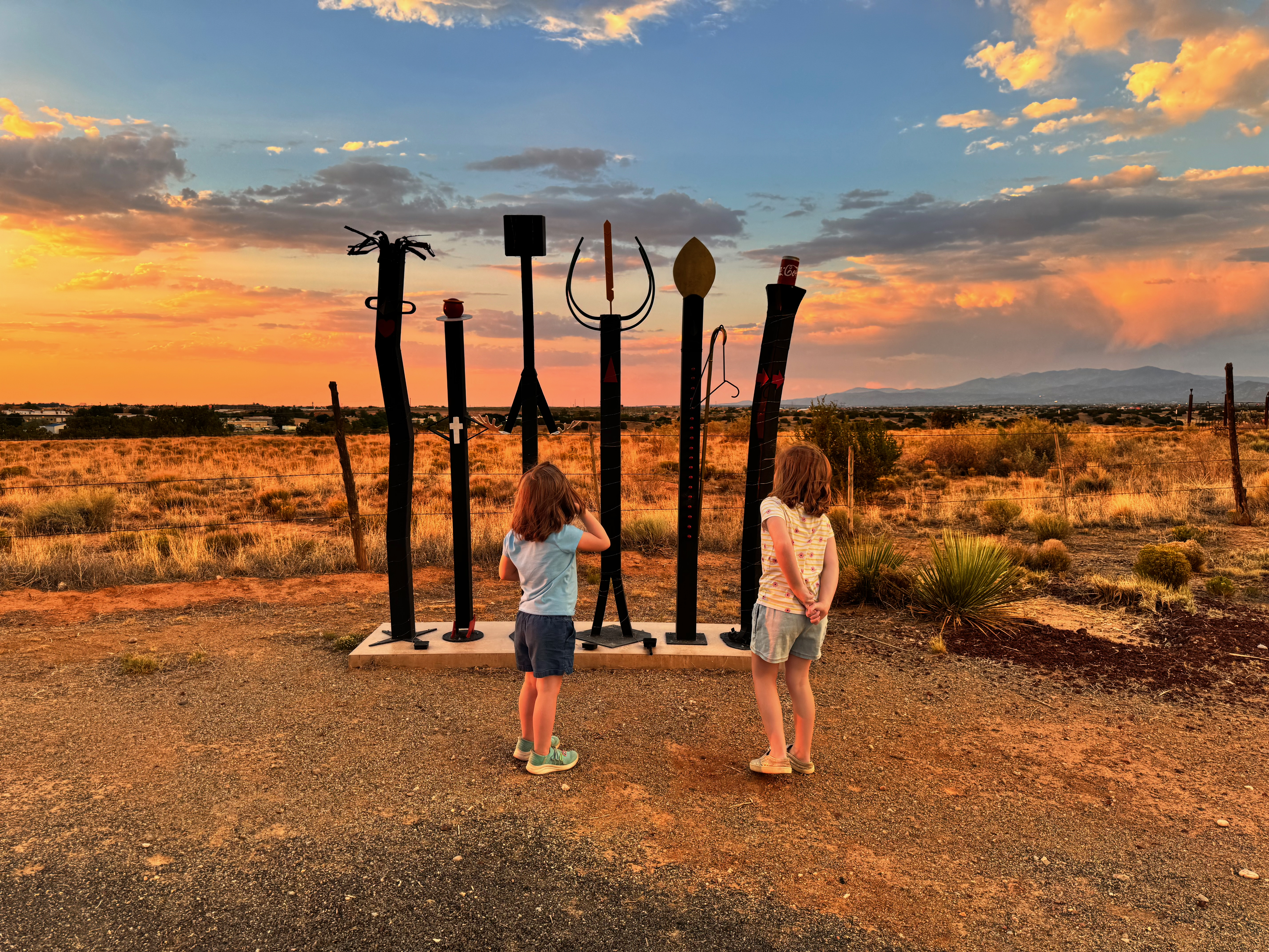 Lillian and Emery standing at the metal sculptures at Santa Fe Skies, watching the sun go down over the high desert