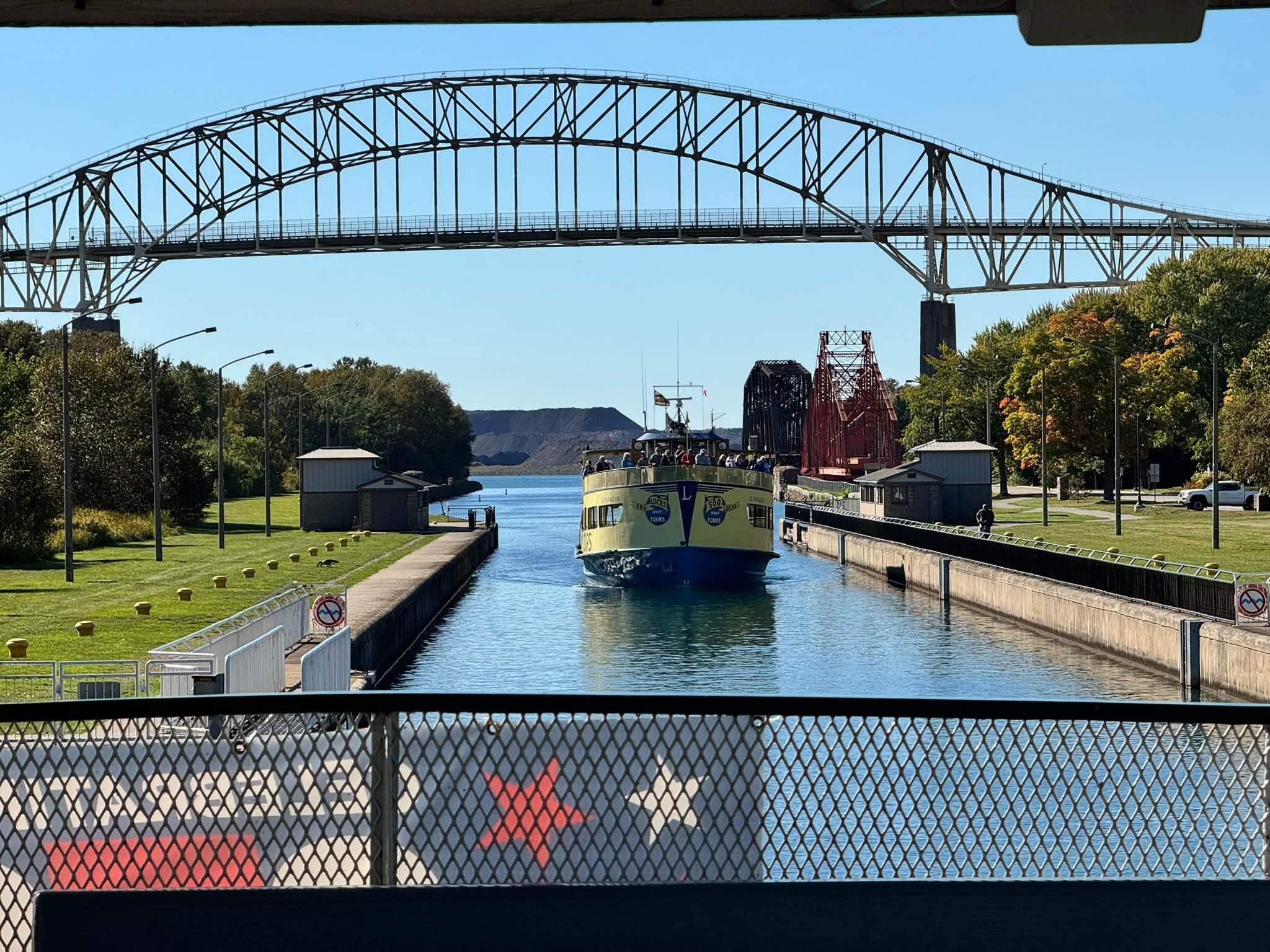 A yellow and blue Soo Locks tour boat heading toward us through the lock channel, the steel arch of the International Bridge overhead and early fall color along the banks