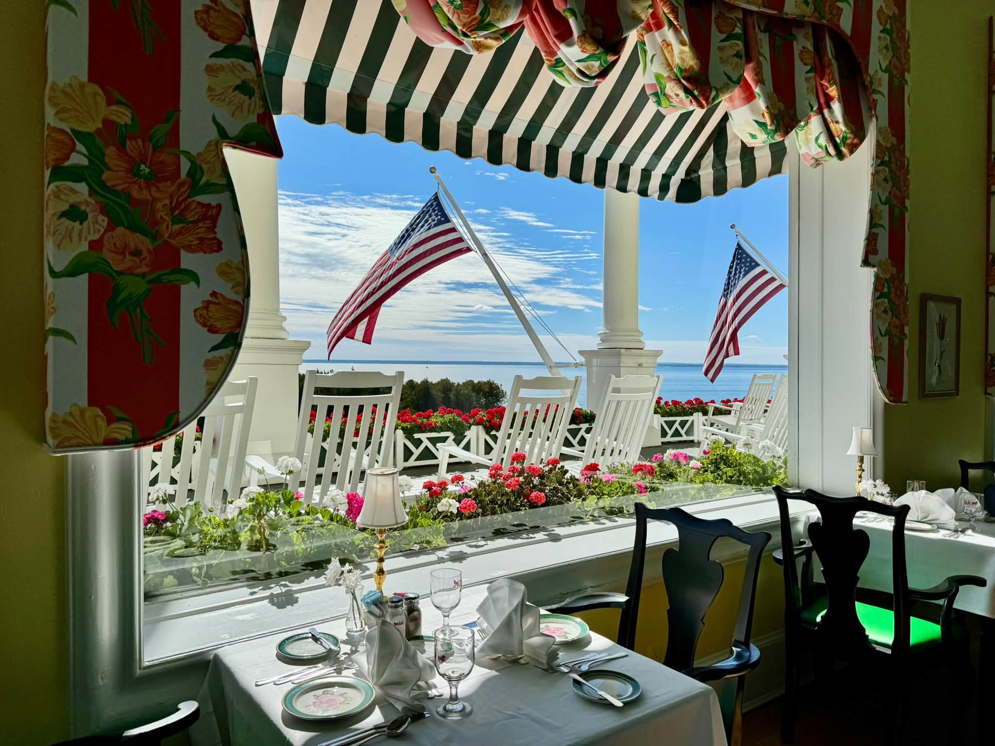 A white-clothed table set for lunch beside a tall window framed by floral drapes and a striped valance, looking out on a porch of white rocking chairs, red geraniums, American flags, and the blue straits beyond