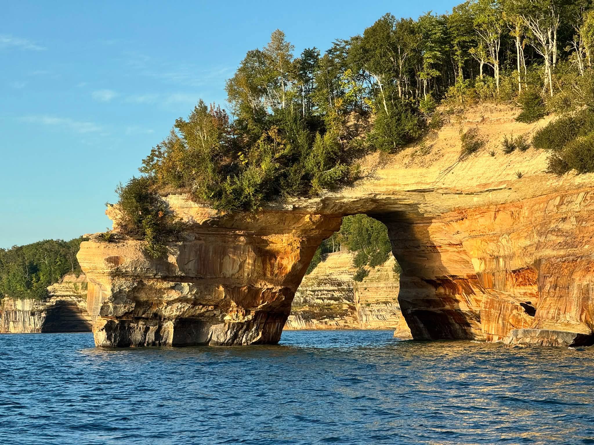 A sandstone arch streaked orange, tan, and rust glowing in low evening sun at Pictured Rocks, trees growing on top and Lake Superior rippling below