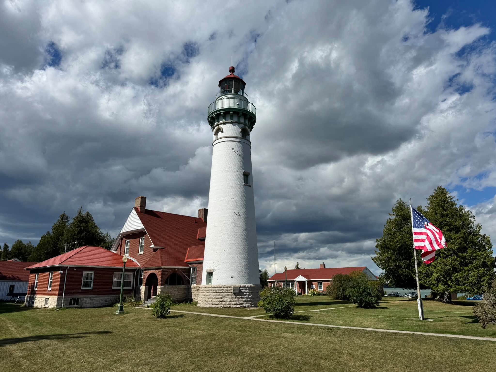 A tall white lighthouse tower with a green lantern gallery beside a red-brick keeper’s house with a red roof, an American flag flying under dramatic clouds