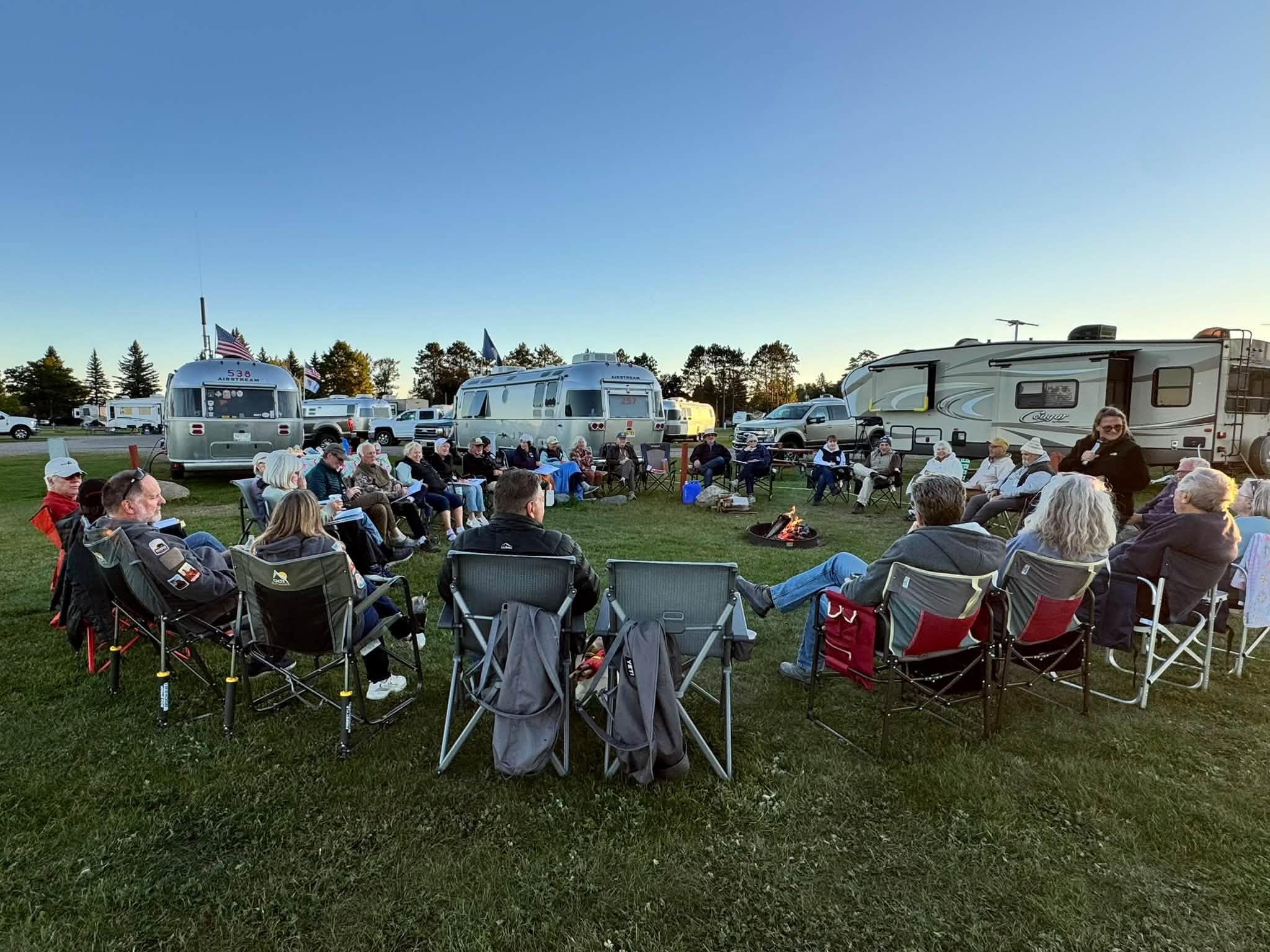 Caravan members in camp chairs gathered in a wide circle around a small campfire at dusk, Airstreams and a fifth-wheel parked behind them under a clear sky