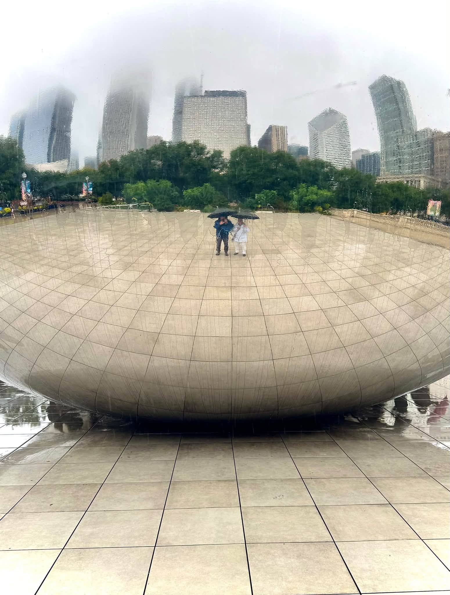 Tom and Sandra under umbrellas, reflected in the curved mirrored surface of Cloud Gate, with fog-shrouded Chicago skyscrapers rising behind