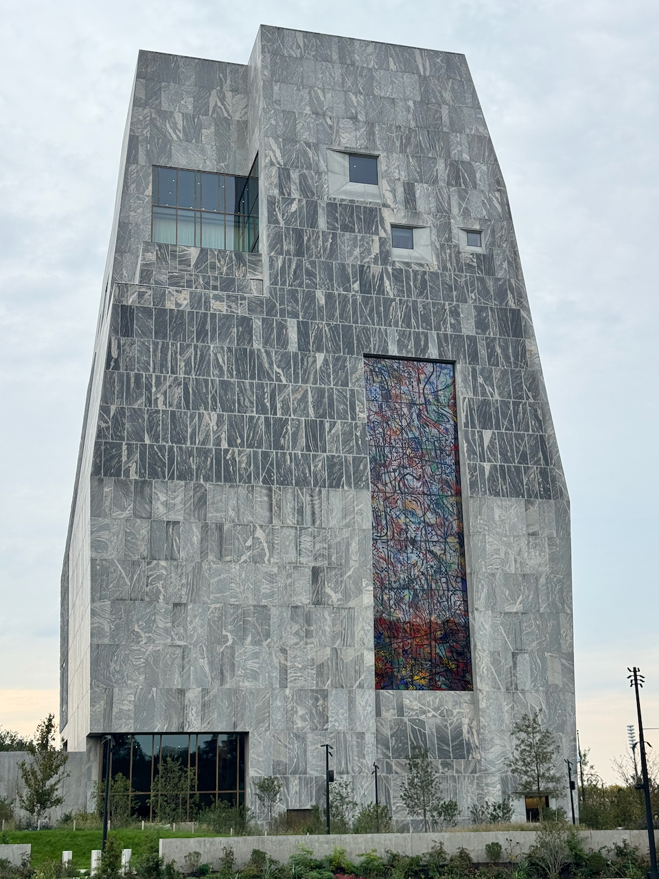 The Obama Presidential Center museum tower, clad in gray veined stone, with a tall multicolored stained-glass-style panel set into its face under an overcast morning sky