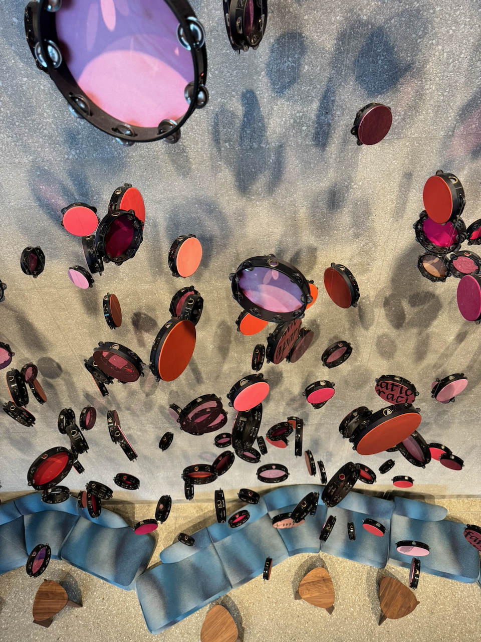 Looking up at dozens of tambourines with red, pink, and purple heads suspended from a silver ceiling above curving blue lounge seating