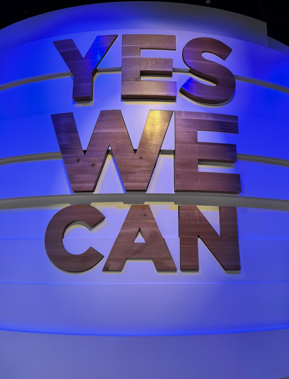 Large wooden letters spelling YES WE CAN mounted on curved white bands glowing in blue light