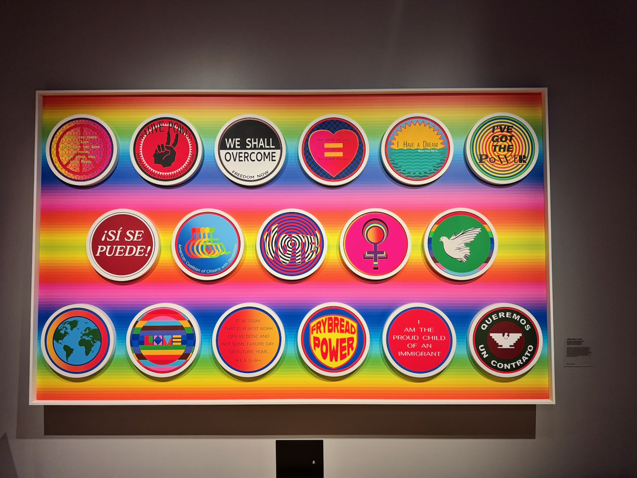 A large framed artwork of eighteen round protest-style buttons — We Shall Overcome, I Have a Dream, Sí Se Puede, Frybread Power, and others — set against bright rainbow stripes