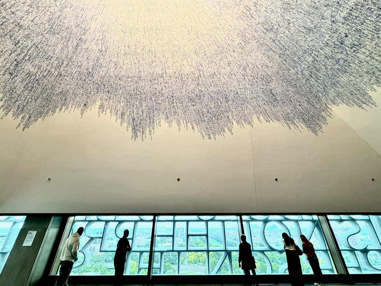A vast cloud of handwritten script spreading across a white ceiling above a band of windows, with visitors silhouetted against the lettered glass