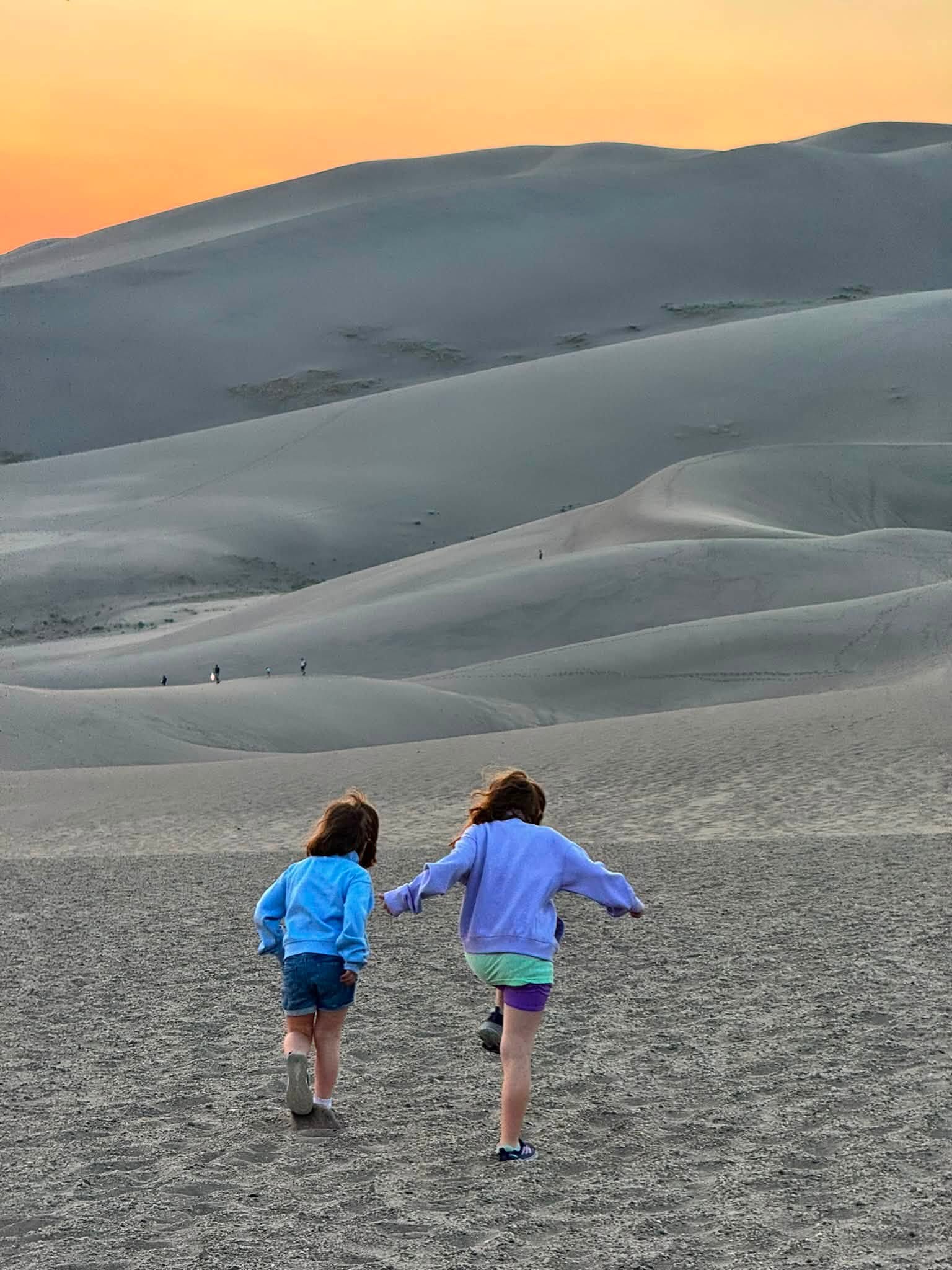 Lillian and Emery running across the sand toward the dunes as the sky turns orange behind them
