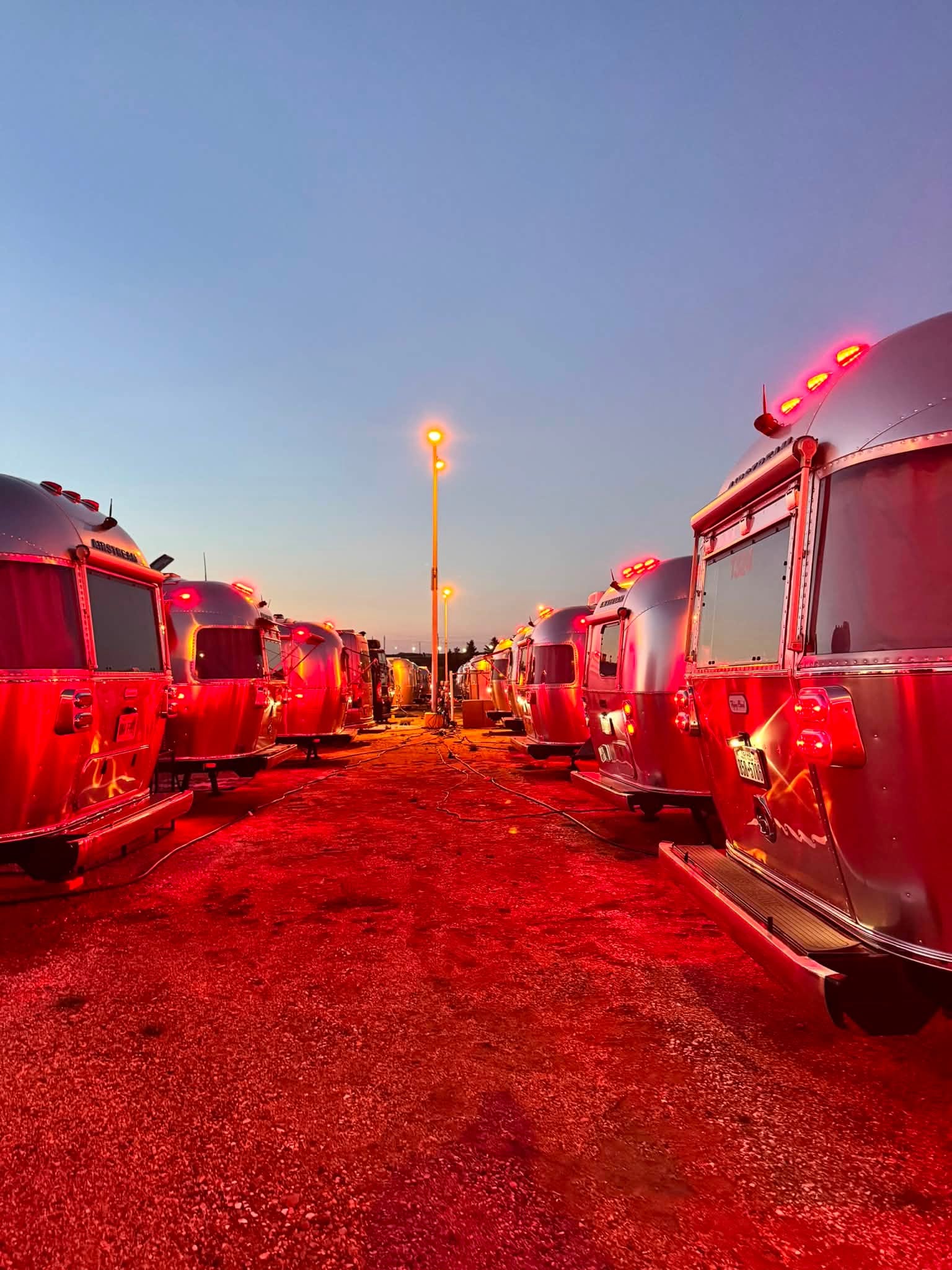 Rows of polished Airstreams at dusk, their red running lights washing the gravel between them in color under a deep blue sky