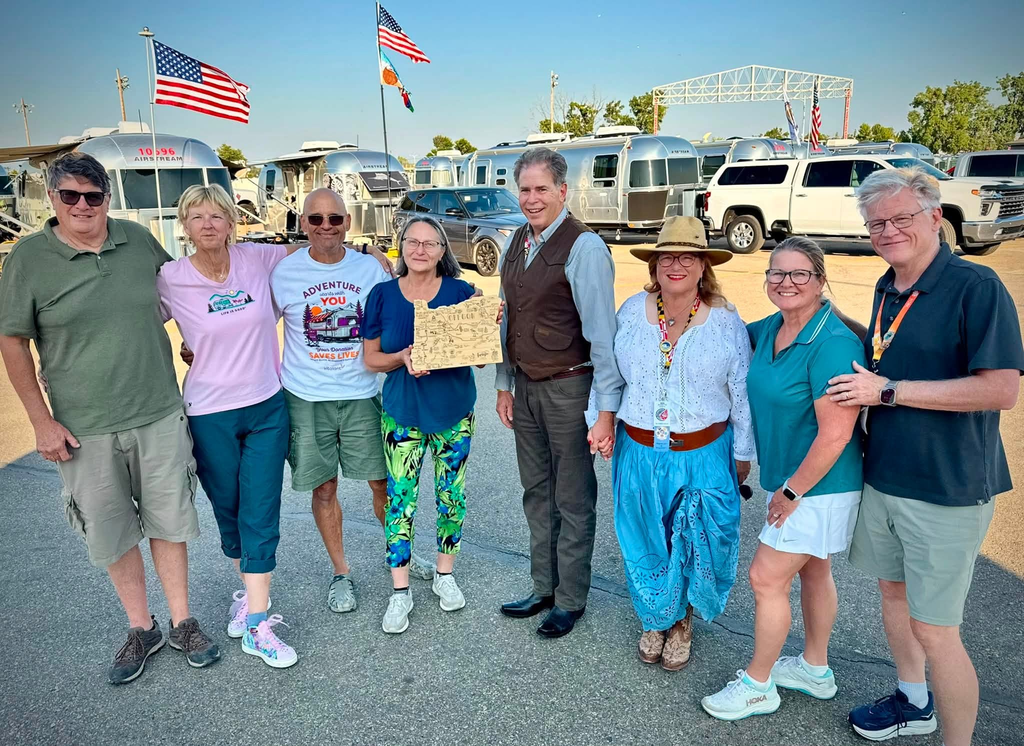 Eight friends from the ExplOregon Caravan gathered between rows of Airstreams under flags, one holding a carved wooden map of Oregon