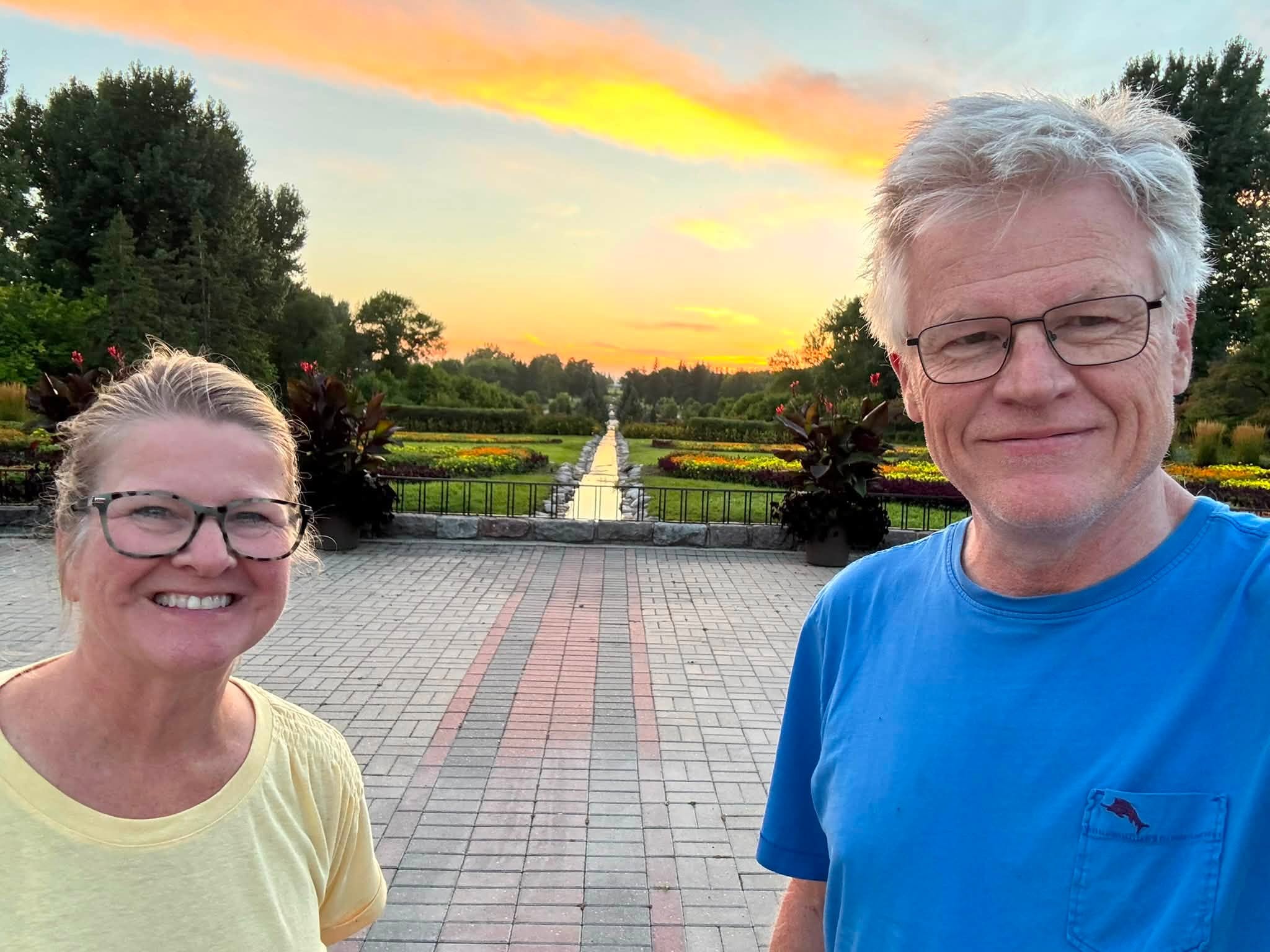 Tom and Sandra side by side in the International Peace Garden at sunset, the formal flower beds and a long reflecting channel running to the horizon behind them, the international boundary passing between them