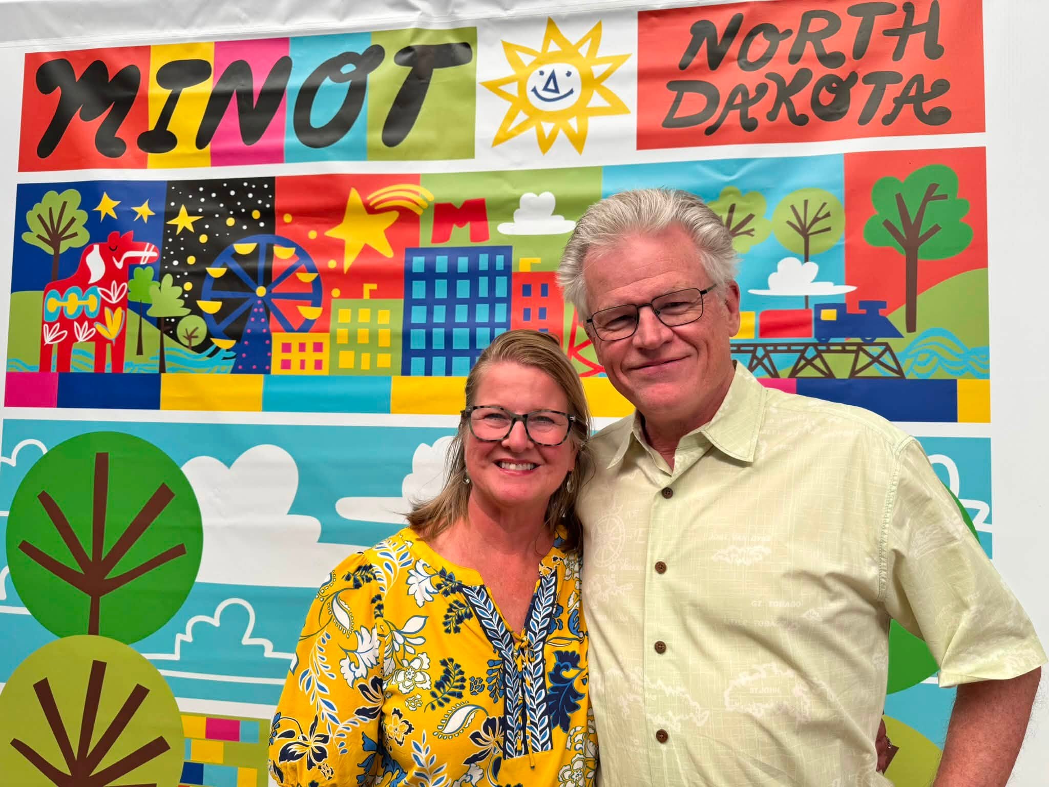 Tom and Sandra in front of a bright illustrated Minot, North Dakota backdrop of trees, buildings, a Ferris wheel and a smiling sun