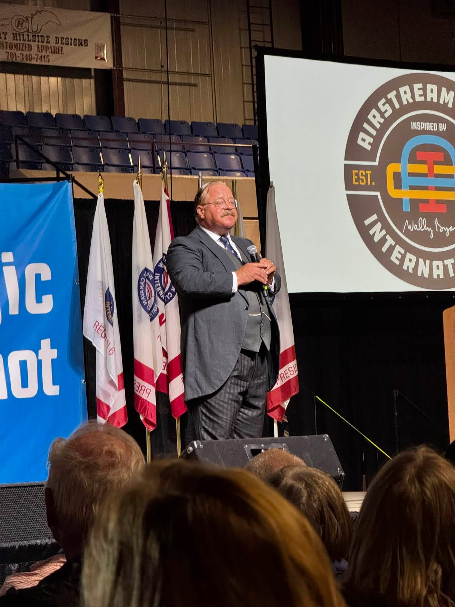 A performer in period suit and vest portraying Theodore Roosevelt, speaking into a microphone on the rally stage beside the Airstream International logo and a row of club flags