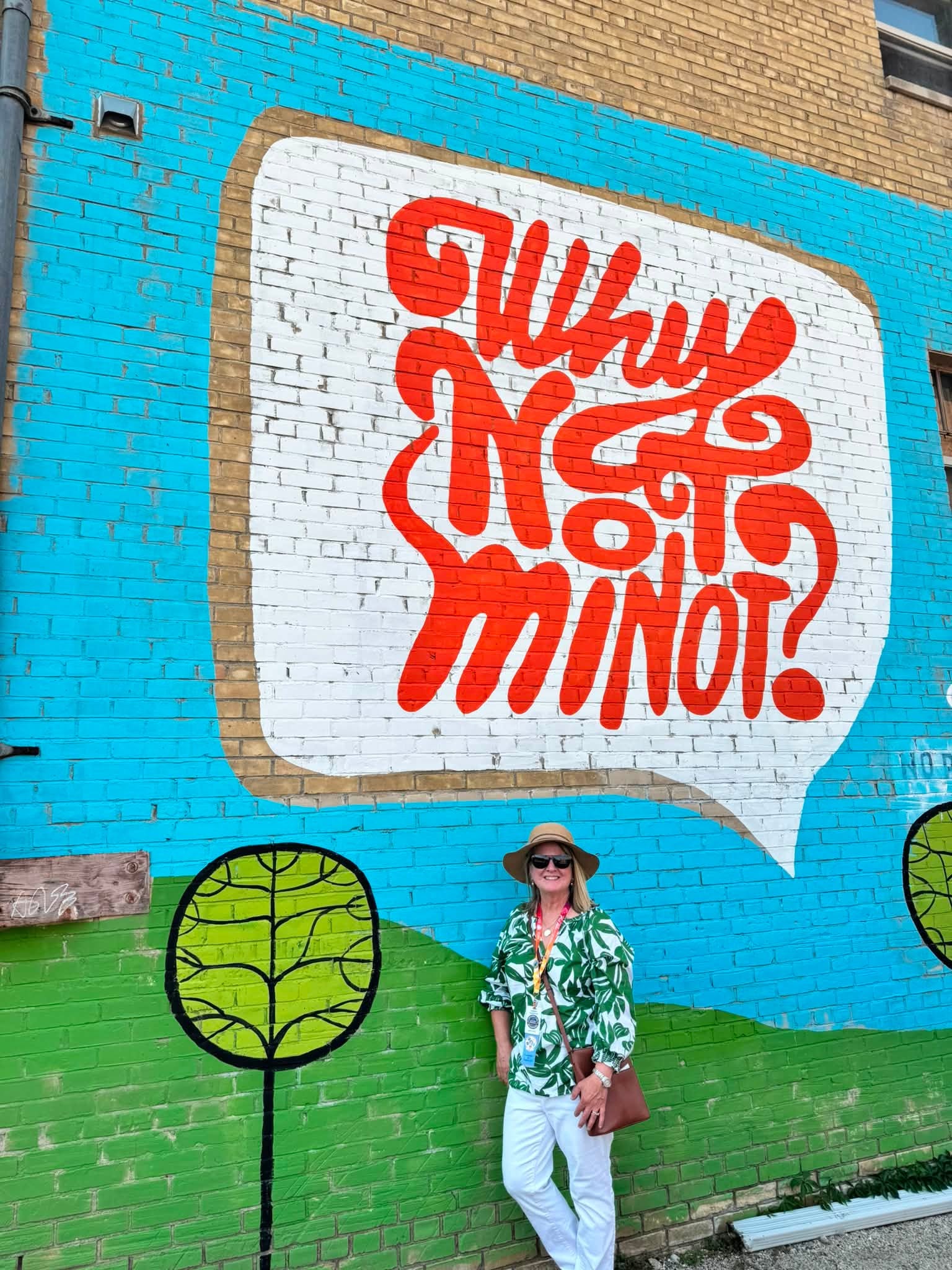 Sandra in a sun hat standing beneath a downtown Minot mural, a giant red speech bubble on brick reading “Why Not Minot?”