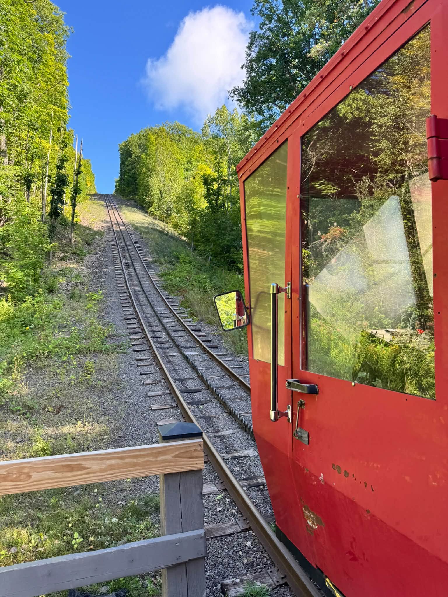 Looking up the steep cog railway incline from beside the red tram car, the single track running straight up a grassy cut between green trees under a blue sky