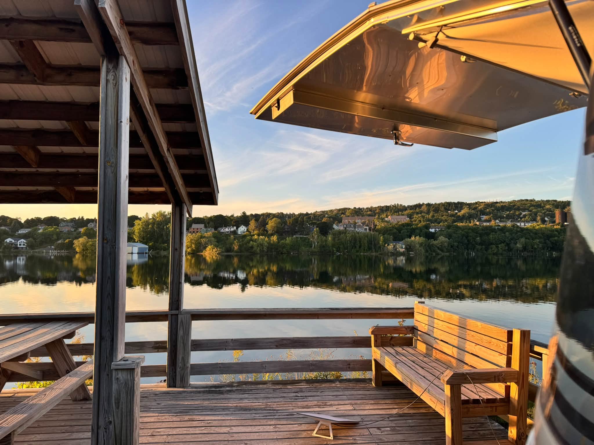 Belle’s awning beside a wooden picnic shelter at Houghton City Park, the waterway glassy in low evening light with the far shore and its houses mirrored in the water