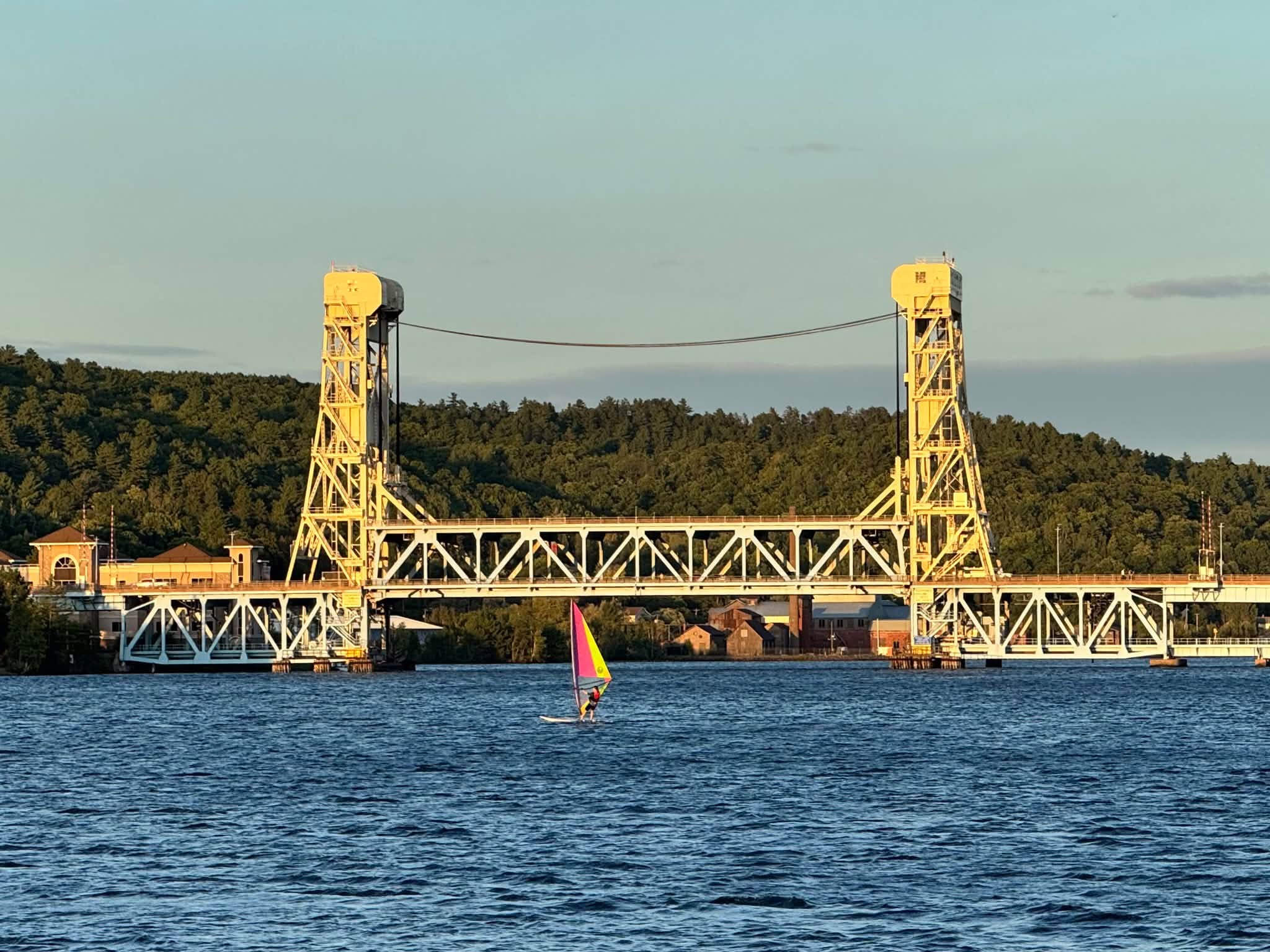 The yellow Houghton–Hancock lift bridge lit by low evening sun, a lone windsurfer with a pink and yellow sail crossing the blue waterway in front of it