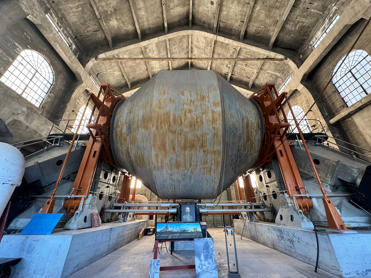 The enormous cable drum of the Quincy Mine hoist filling a tall concrete engine house, braced by orange steel frames with arched windows on either side