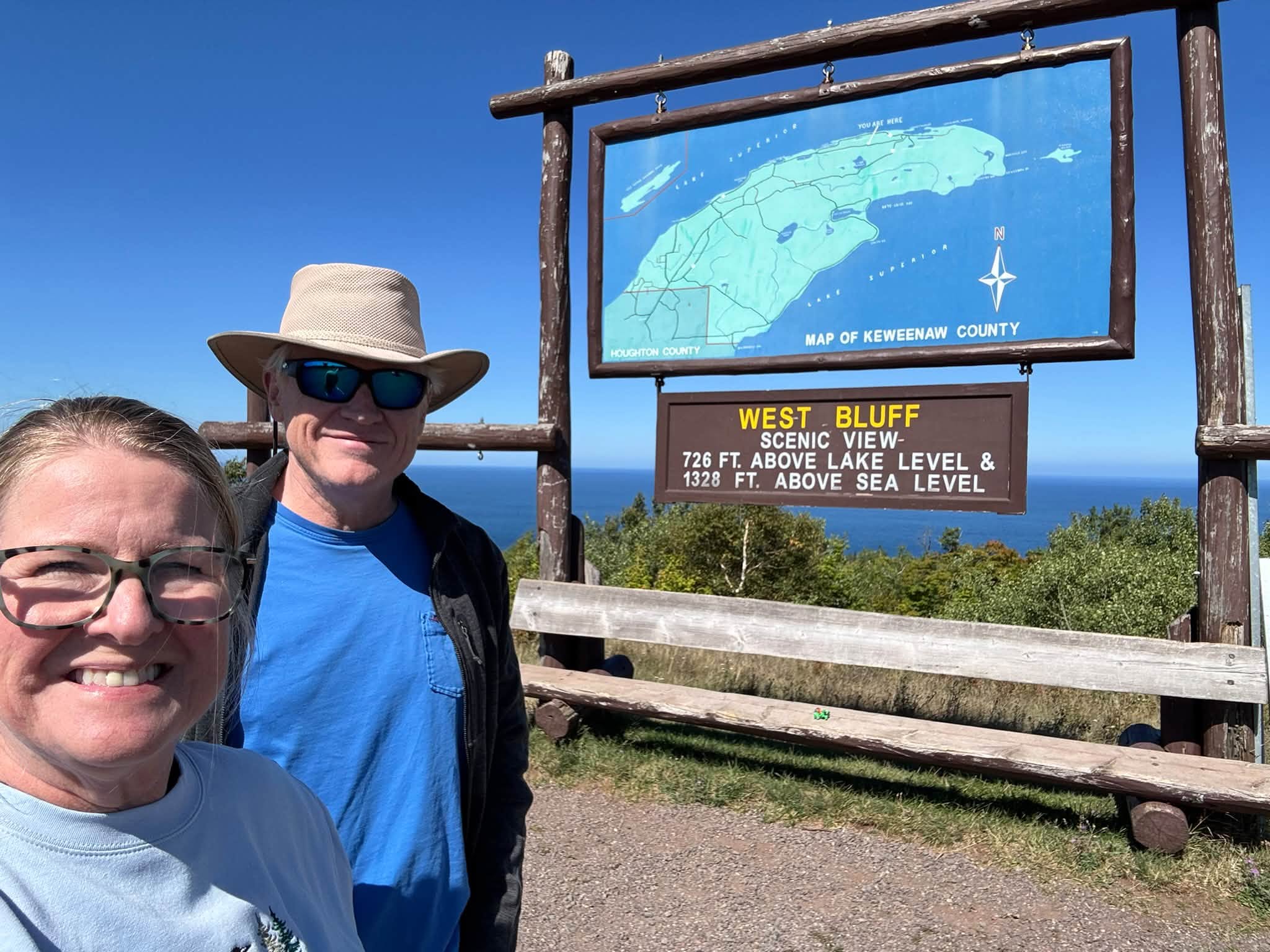 Sandra and Tom smiling in front of a wooden sign reading West Bluff Scenic View, 726 ft above lake level and 1328 ft above sea level, with a map of Keweenaw County above it and Lake Superior stretching out behind