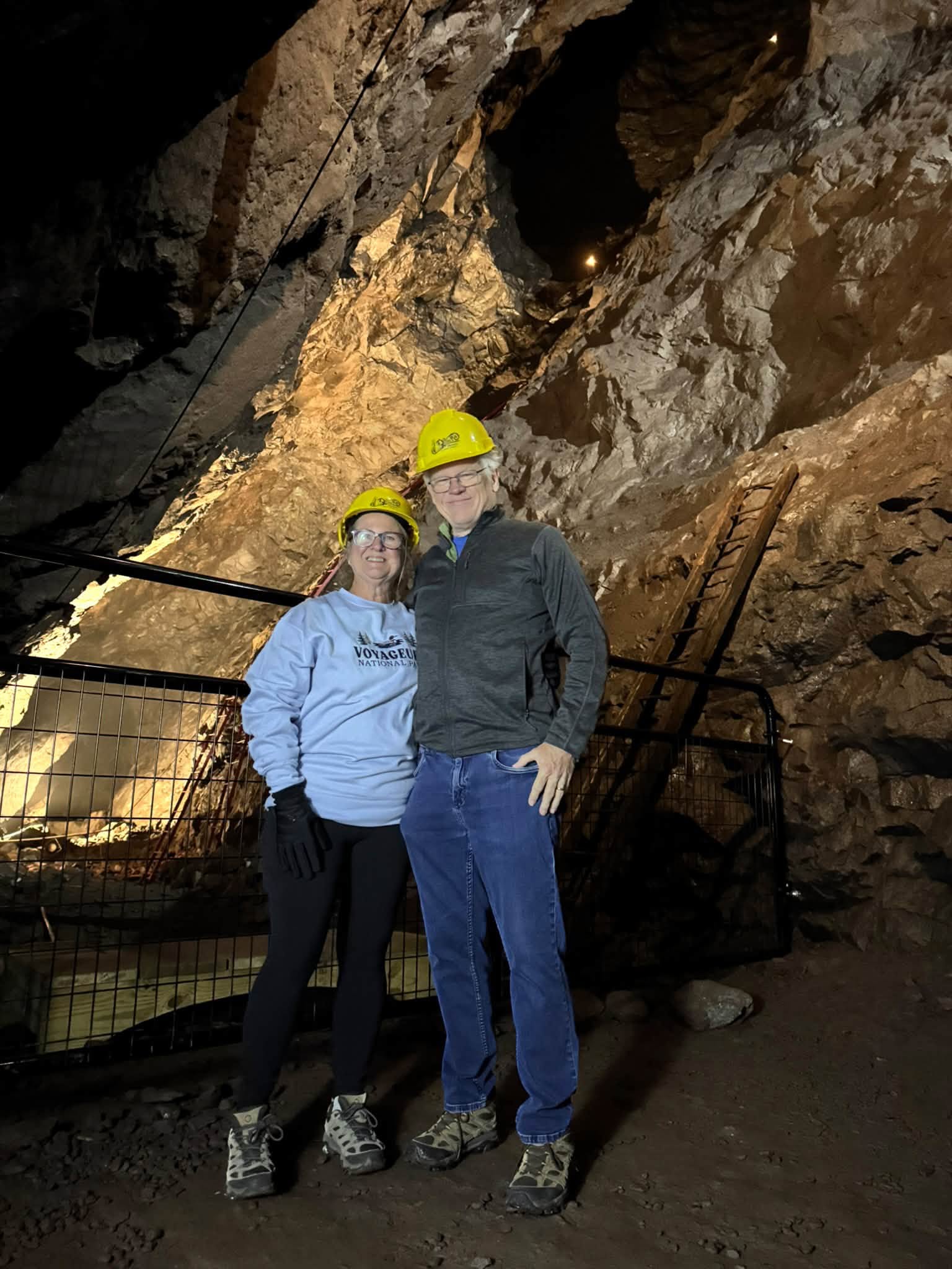 Tom and Sandra in yellow hard hats standing together inside the rough lit rock tunnel of the Quincy Mine