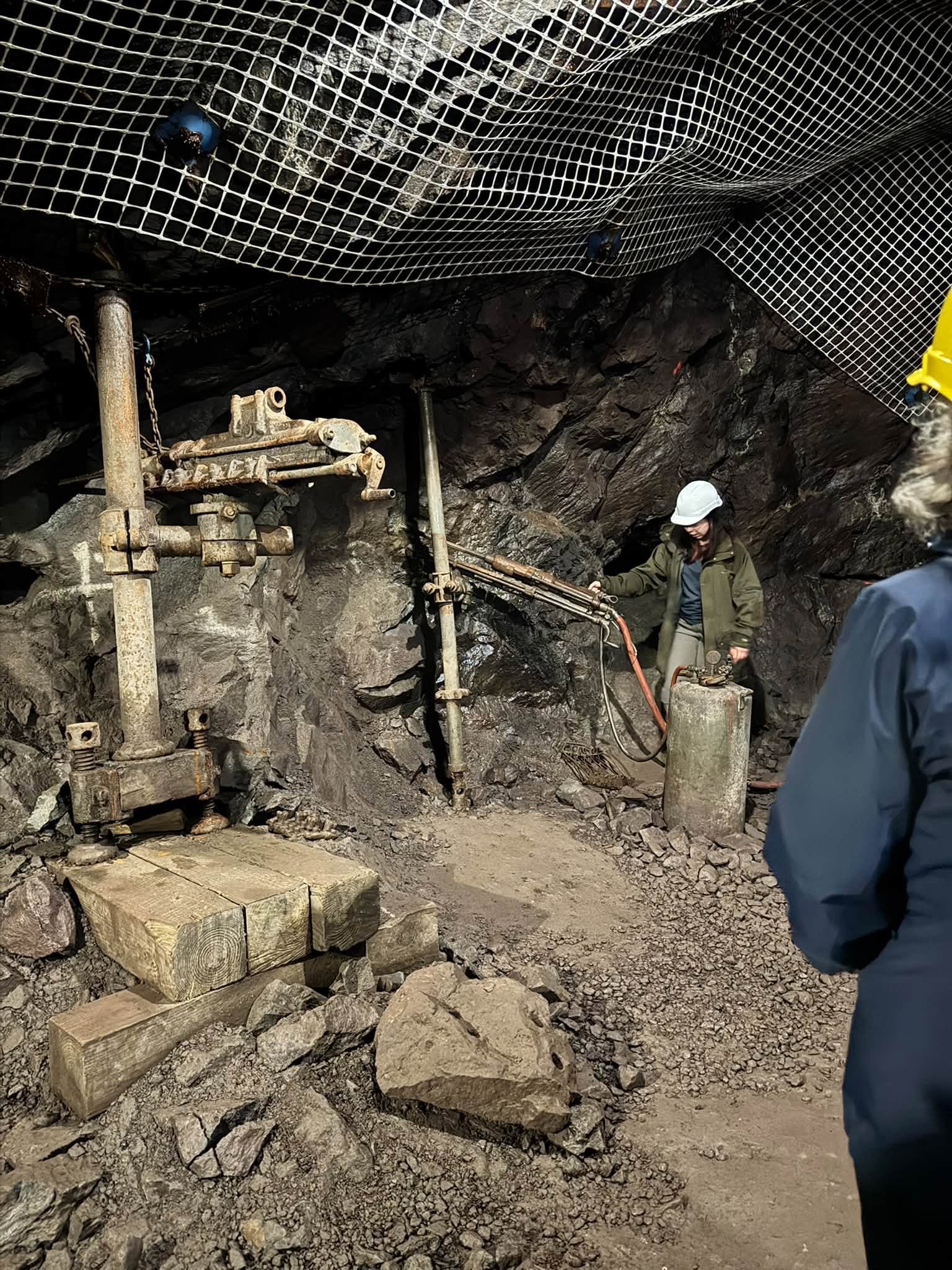 A rusted rock drill mounted on timbers in a mine drift, wire mesh pinned to the ceiling overhead and a guide in a white hard hat demonstrating alongside