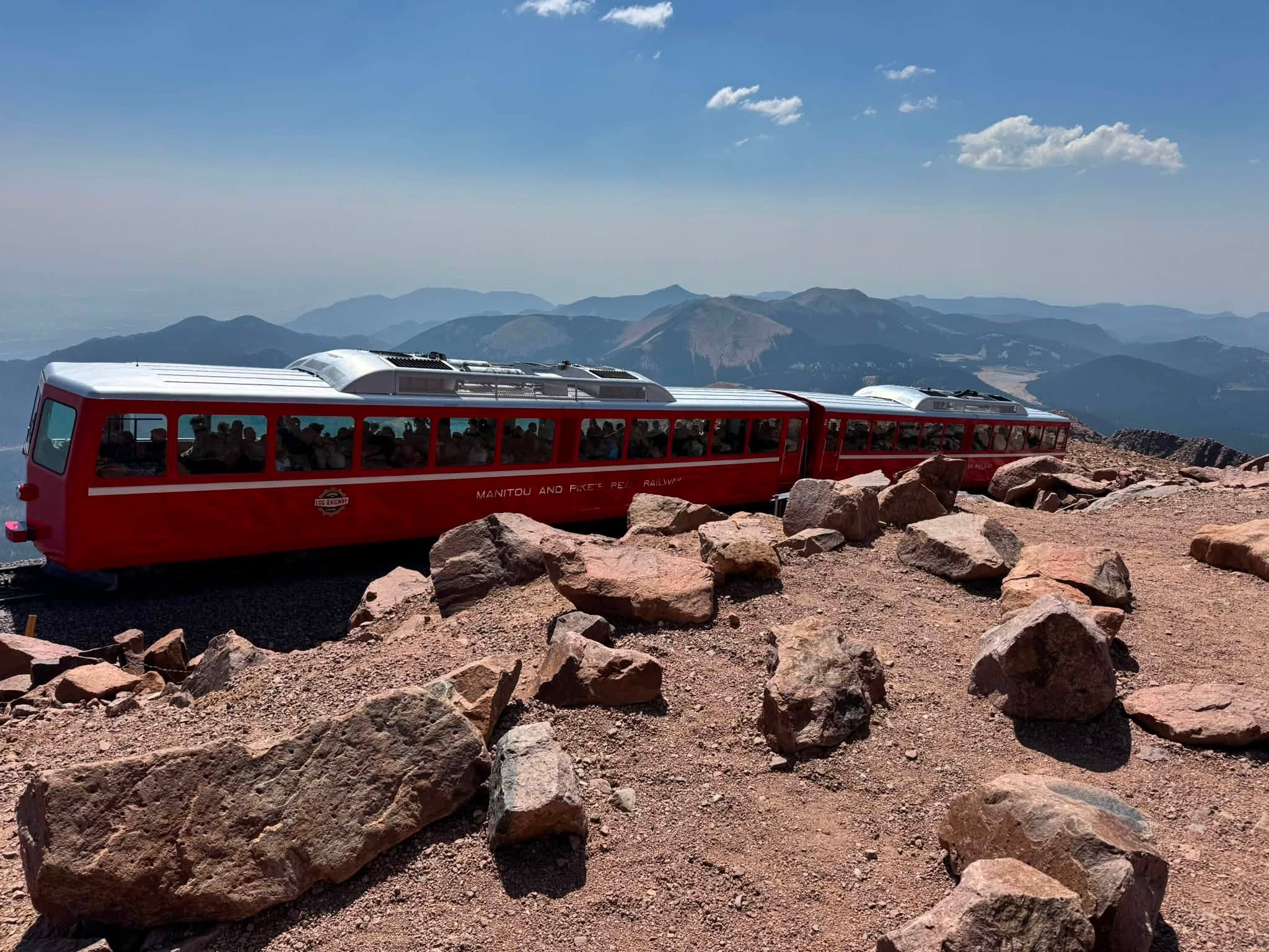 The red Manitou and Pike’s Peak Railway cars parked at the 14,115-foot summit, mountain ranges fading into the haze behind