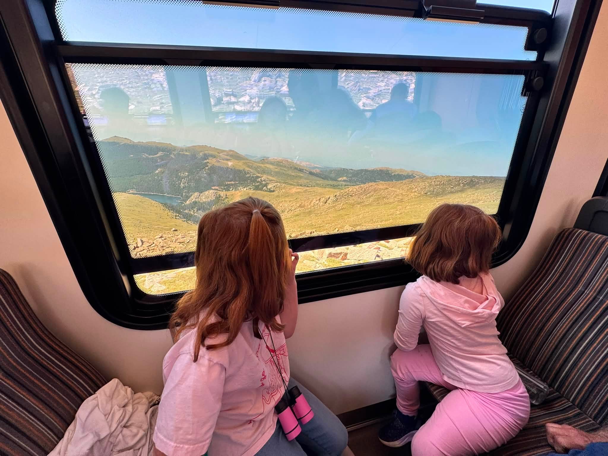 Lillian and Emery kneeling on the train seat, faces to the window, watching the mountains and a reservoir far below