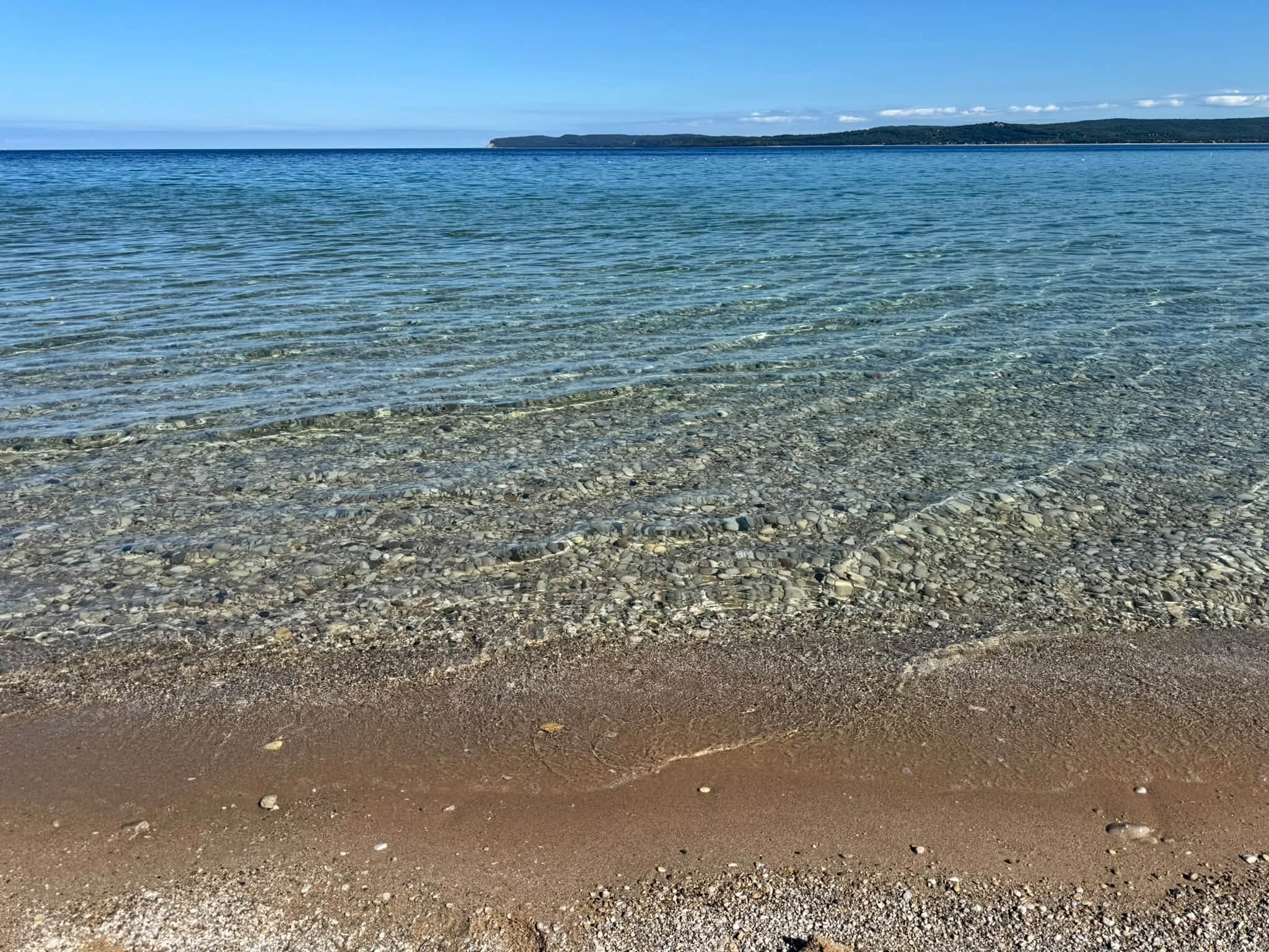 Crystal-clear Lake Michigan water lapping over smooth pebbles onto a sandy beach, a wooded point on the horizon