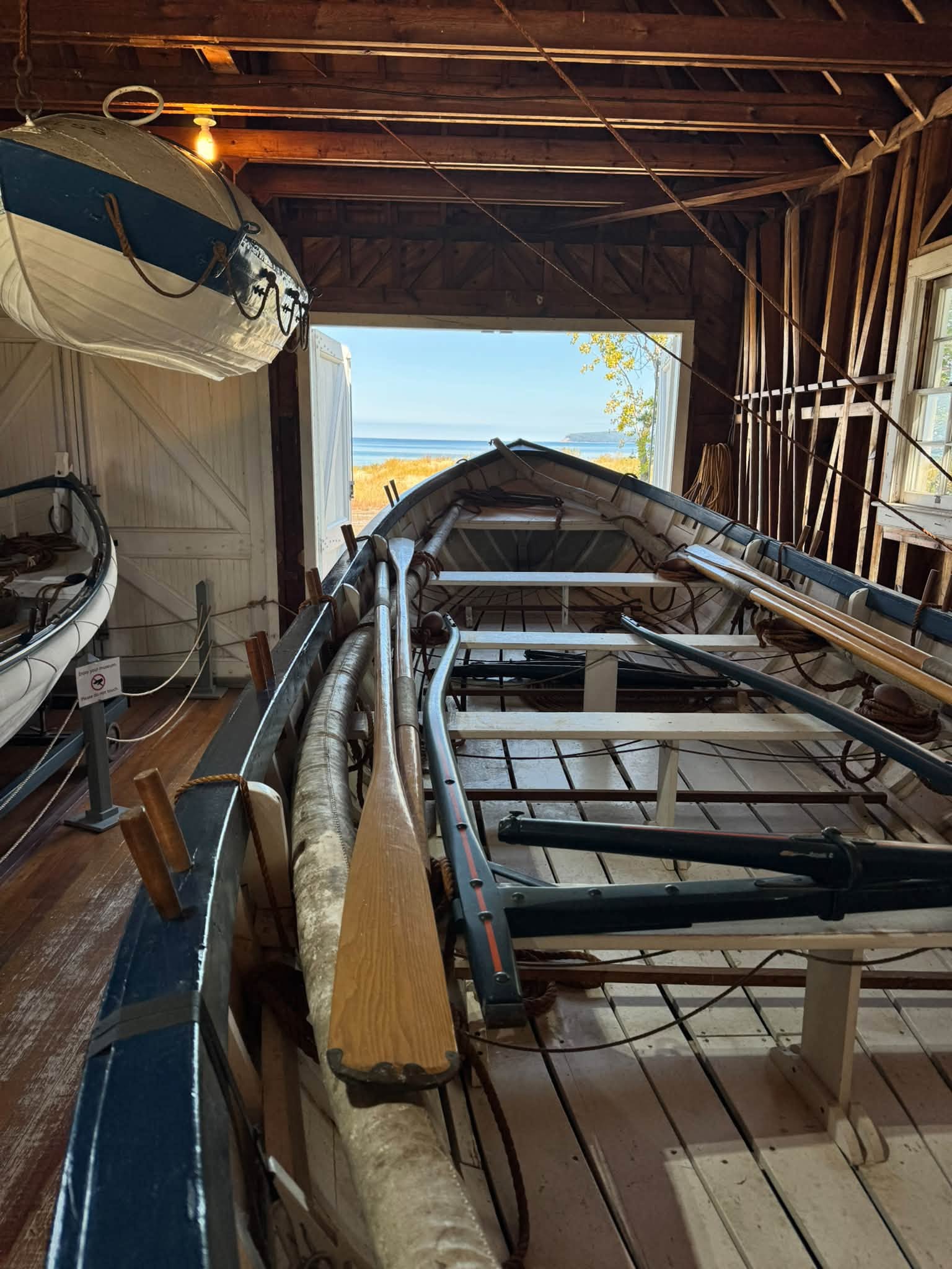 Inside the historic boathouse at the Sleeping Bear Point Life-Saving Station, a long white-and-navy surfboat with oars and coiled line, the open bay door framing the beach and lake