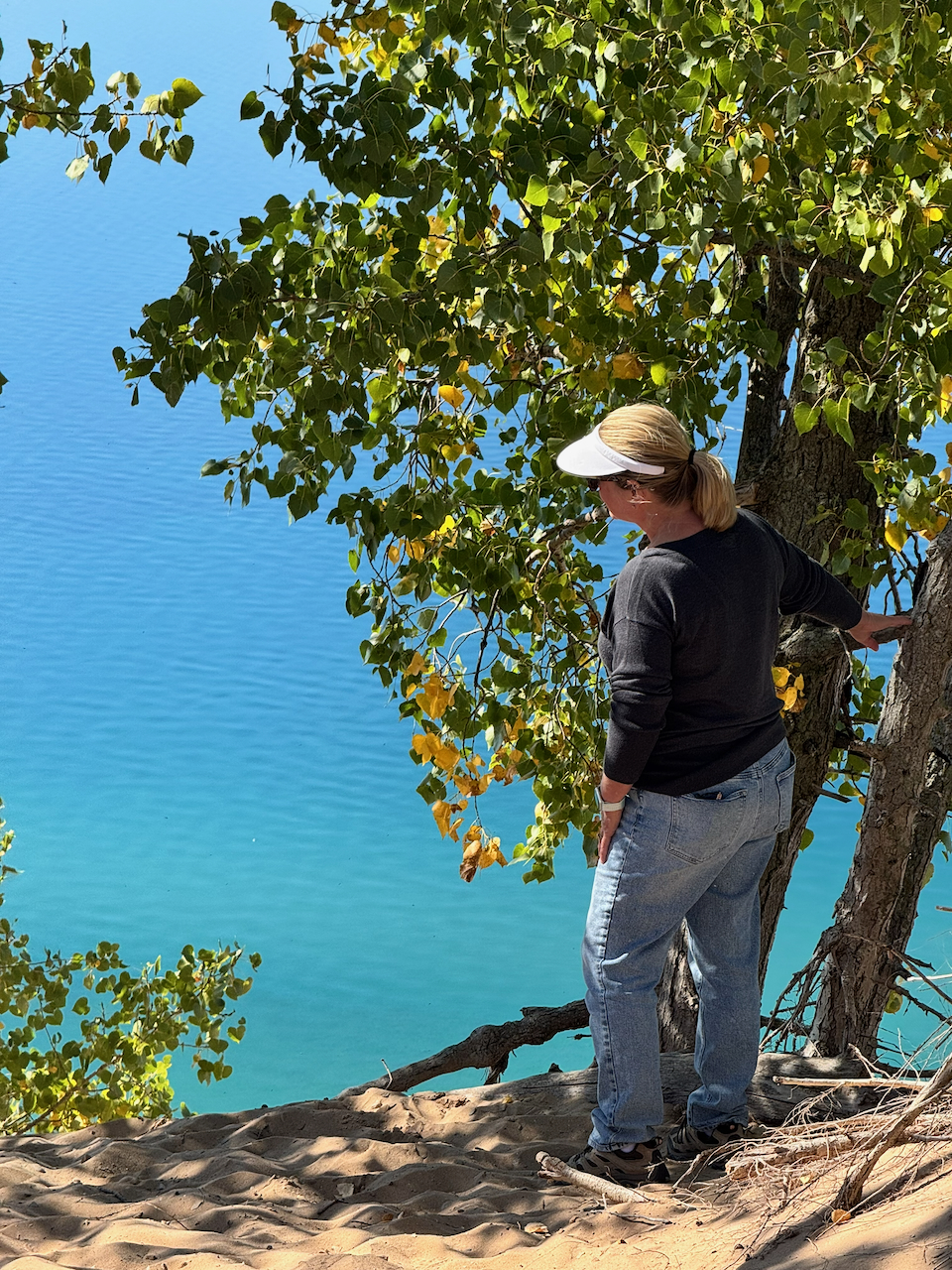 Sandra in a white visor and dark sweater standing at the sandy edge of a dune bluff beside a birch tree, brilliant turquoise water far below