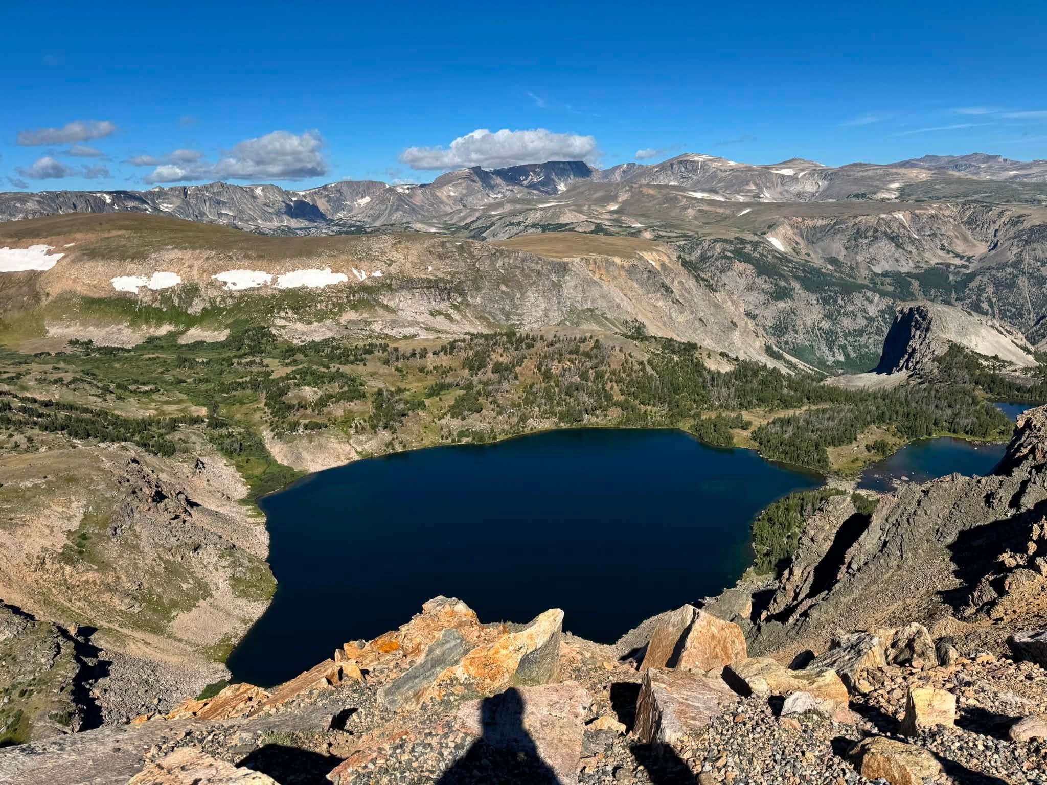 A deep blue alpine lake set in a rocky basin far below the overlook, with snow-streaked peaks running along the horizon