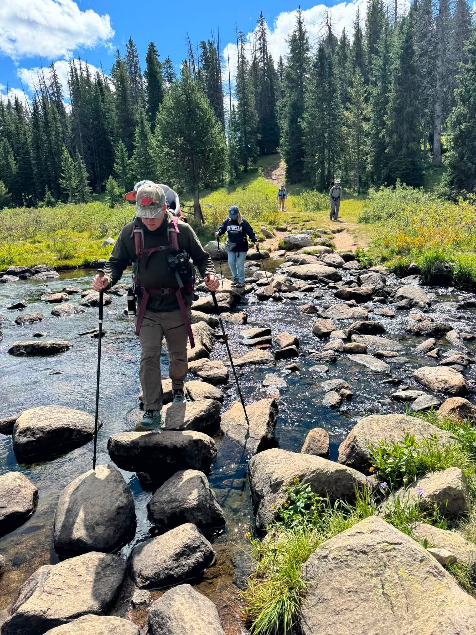 Wes stepping across a rocky alpine stream on stones, trekking poles in both hands and Ben riding in the pack on his back, with the rest of the group following on the trail behind