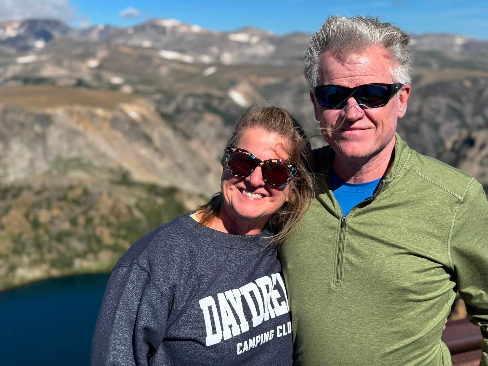 Sandra and Tom standing together at a Beartooth overlook in sunglasses, an alpine lake and snow-dusted mountains behind them