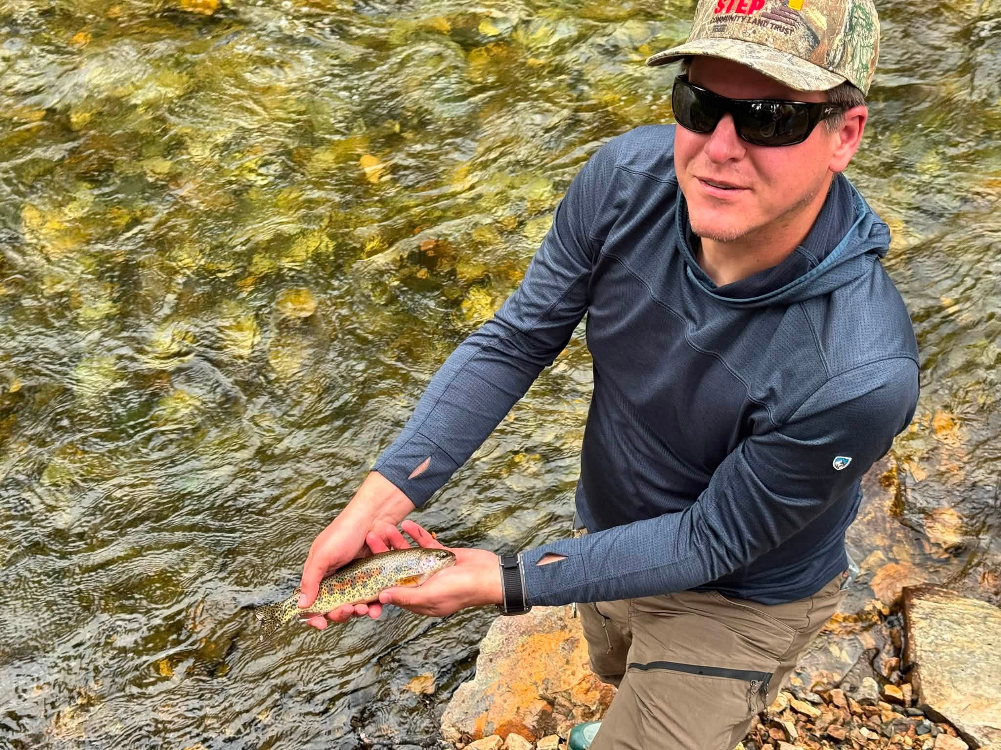 Wes kneeling at the edge of Rock Creek, holding up a small speckled trout over the clear running water