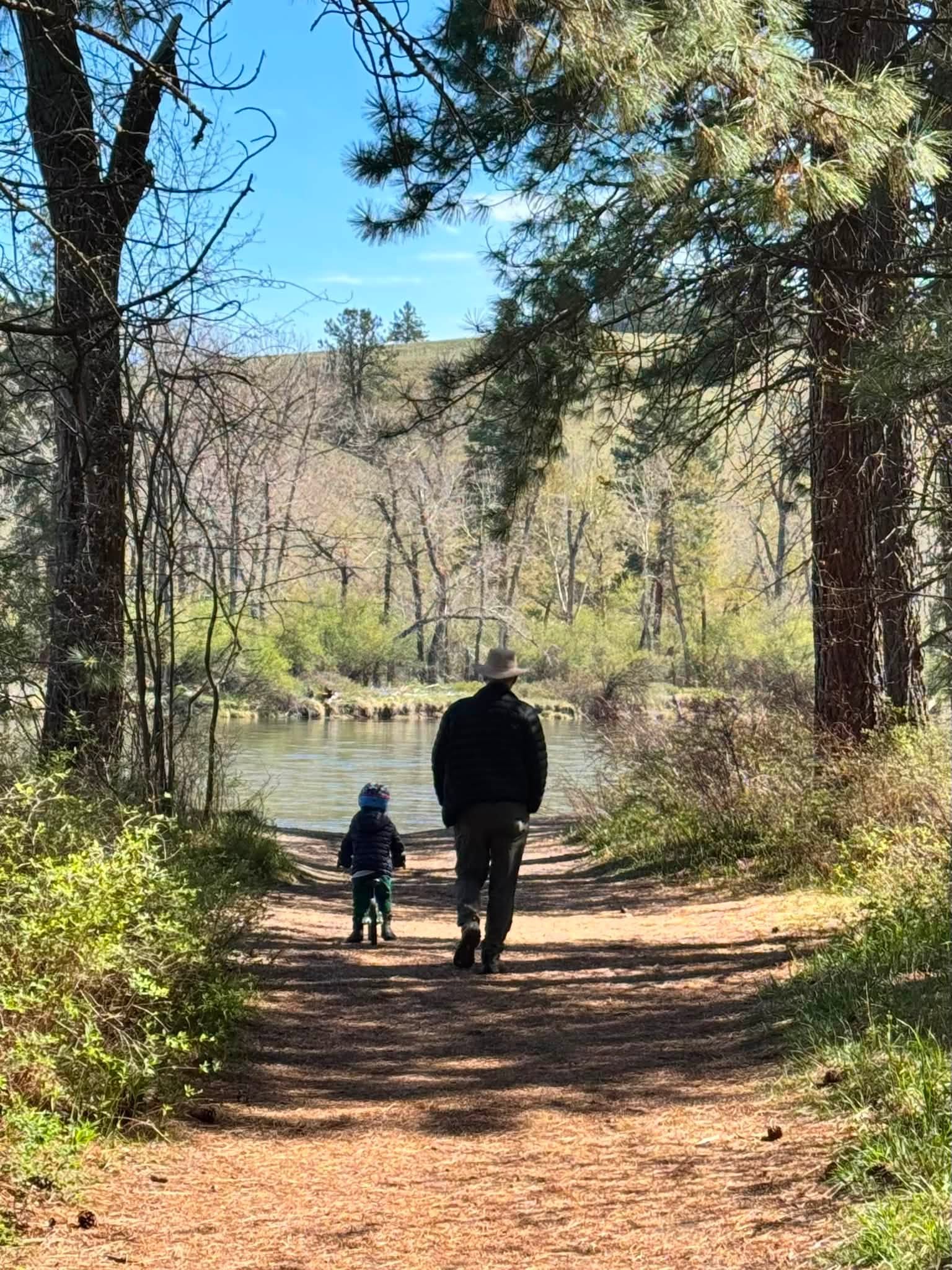 Tom and Bennett walking hand in hand down a shaded trail toward the river in Missoula, Montana