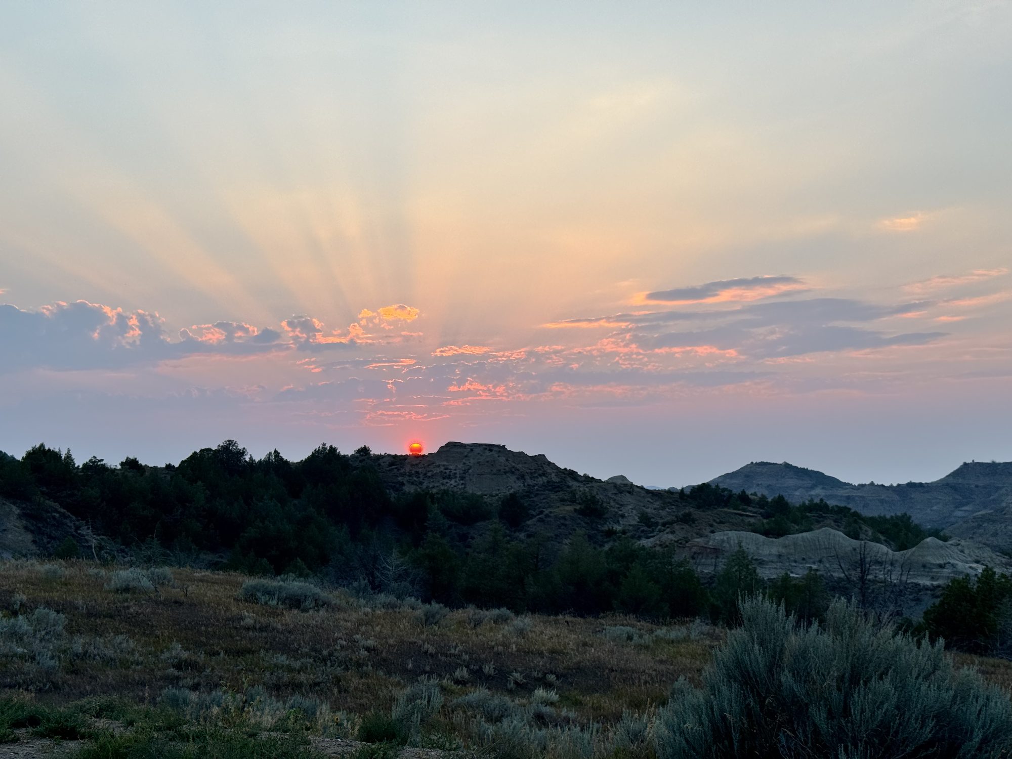 The sun dropping behind a flat-topped butte in Theodore Roosevelt National Park, rays fanning up through pink and gold cloud, sagebrush in the foreground