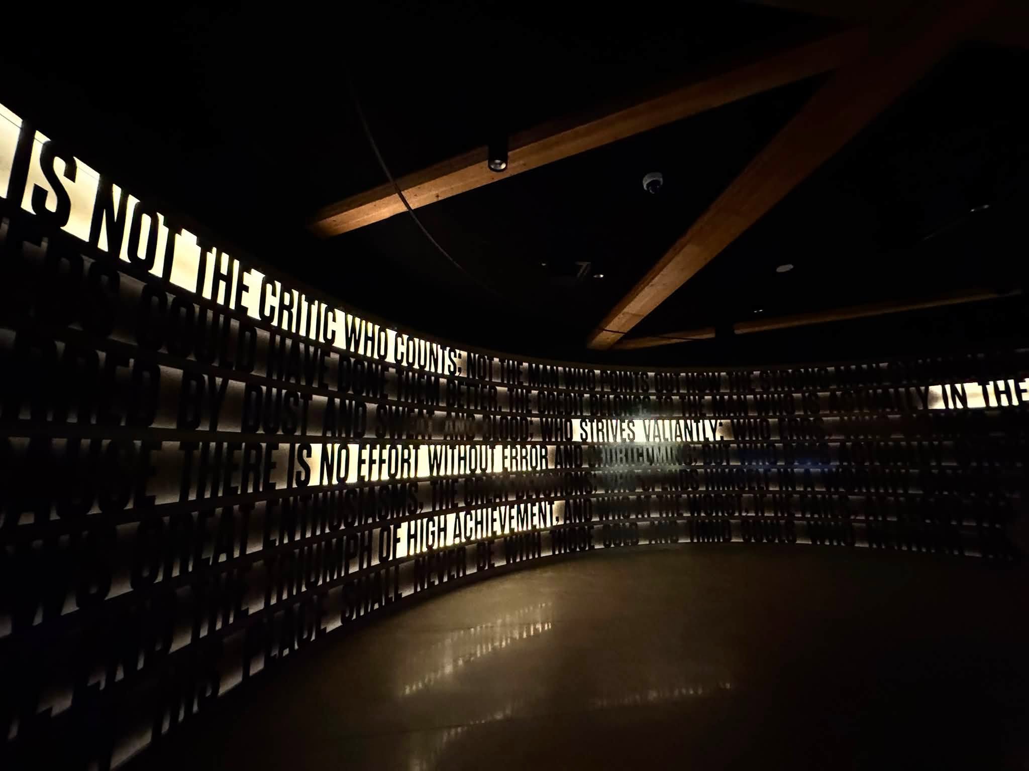 A darkened curved gallery wall carrying Roosevelt’s “Man in the Arena” speech in raised letters, lit so the words catch the light