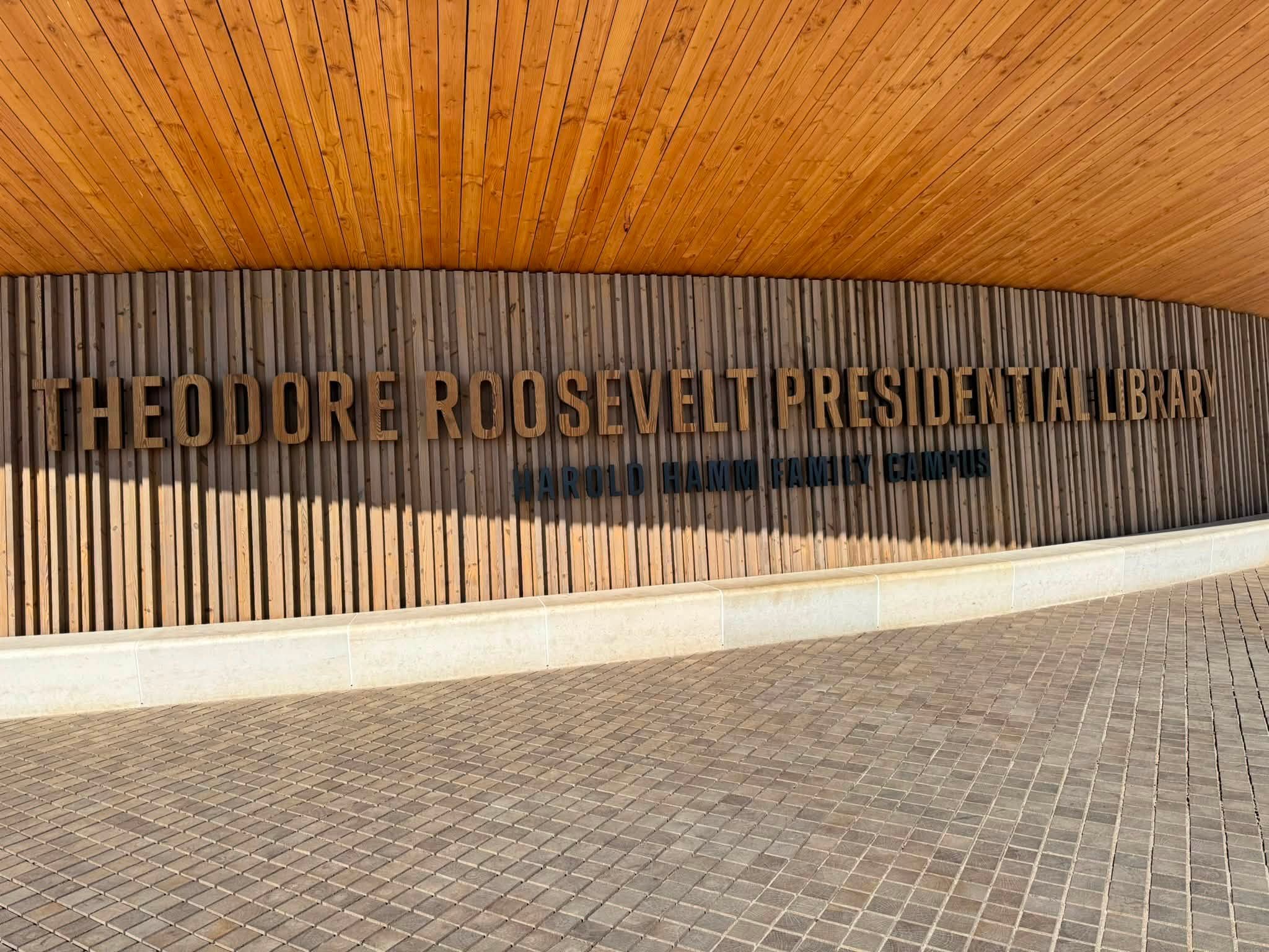 The entrance wall of the Theodore Roosevelt Presidential Library, dark lettering on vertical wood slats beneath a curved timber ceiling