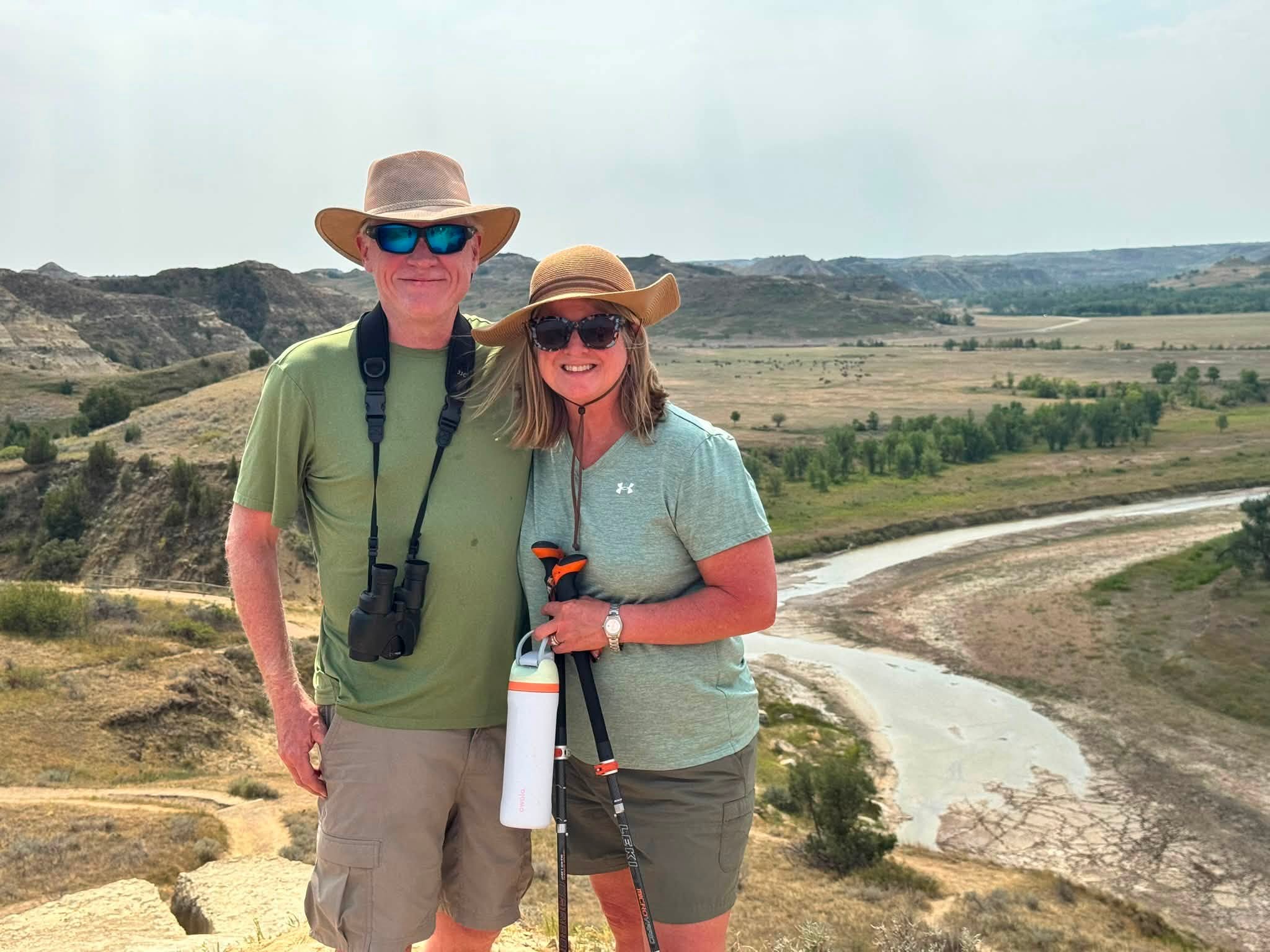 Tom and Sandra in sun hats at an overlook, the Little Missouri River winding through the badlands below and behind them