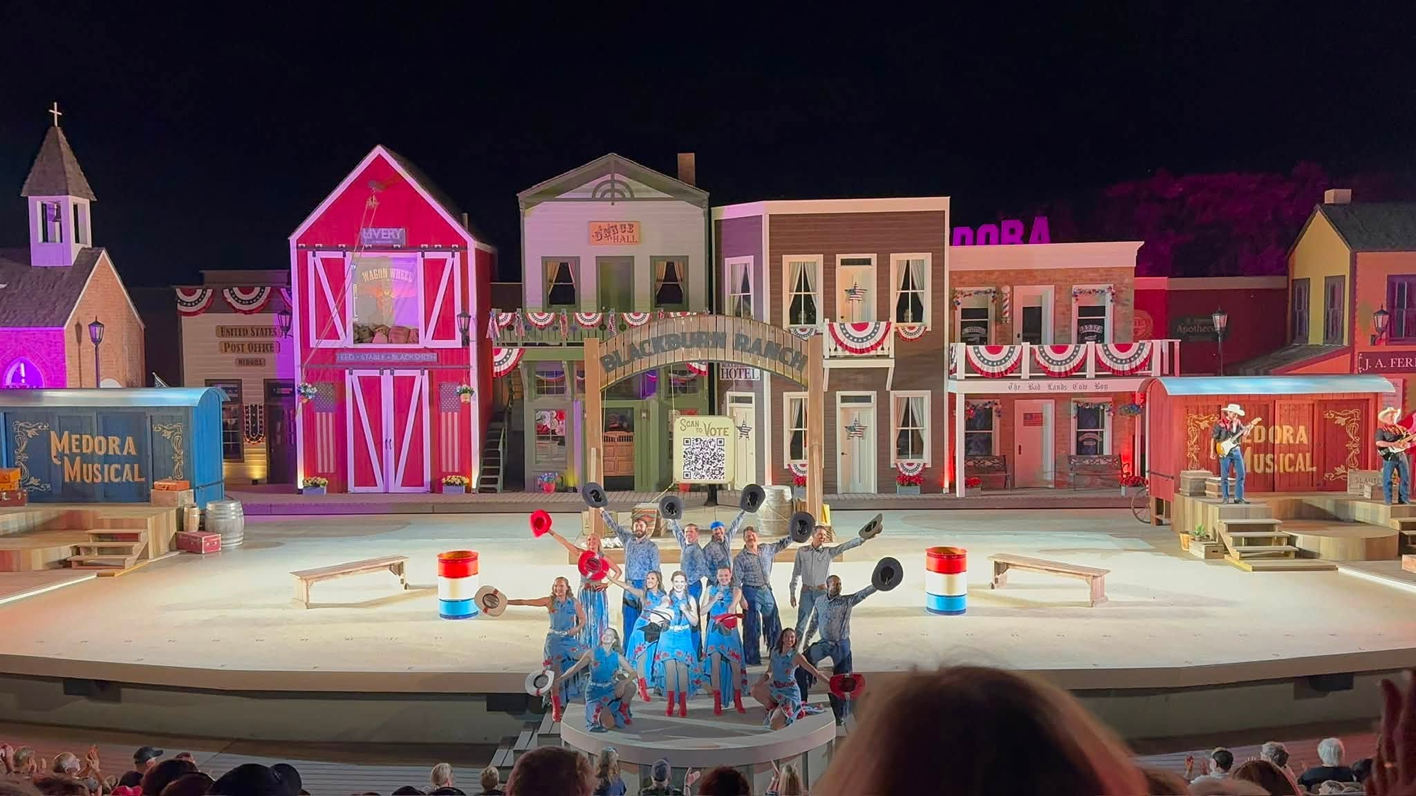 The cast of the Medora Musical in denim and red, white and blue on the open-air stage, backed by a painted Western storefront set under the Blackstone Ranch arch