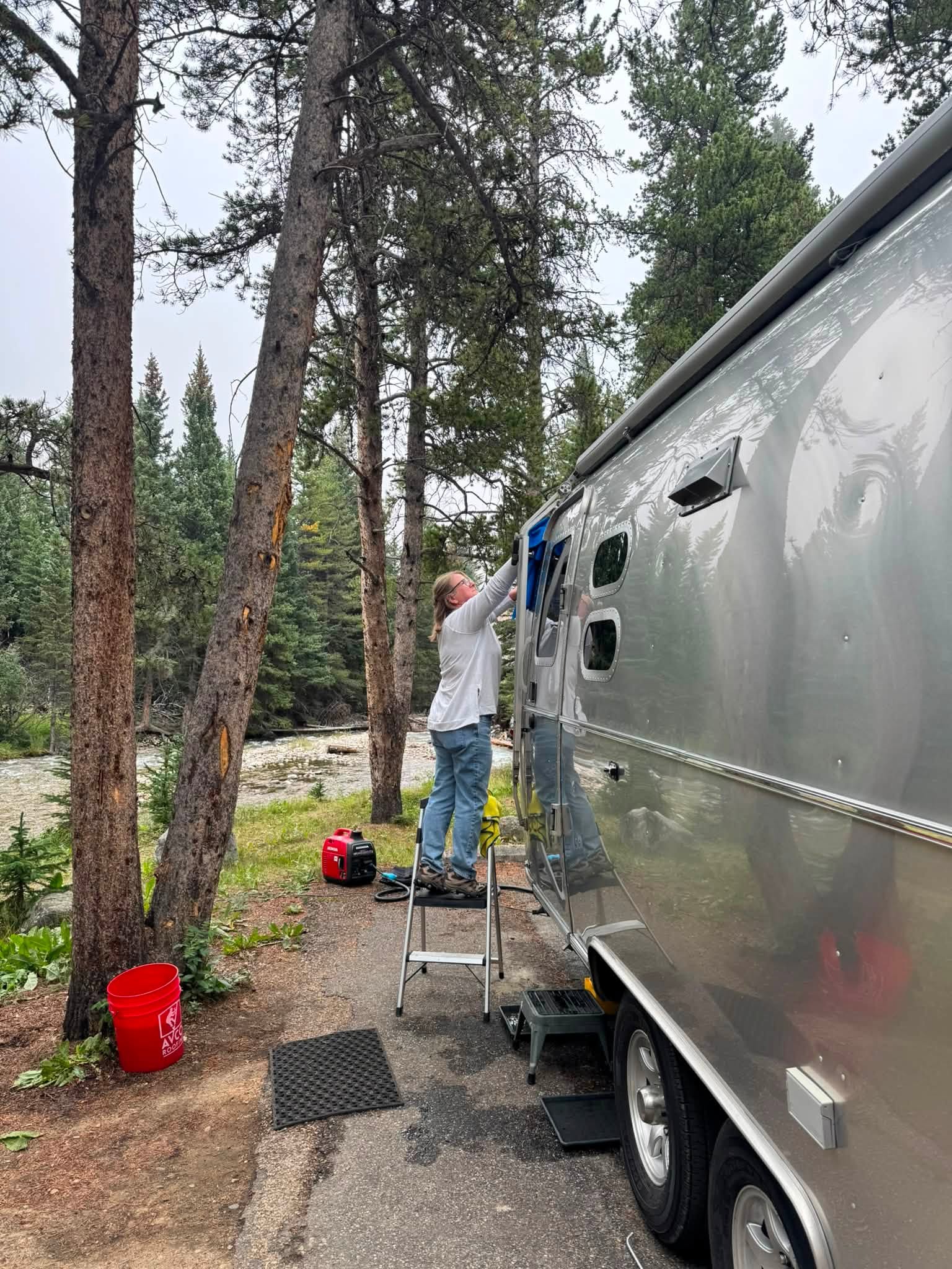 Sandra on a step stool drying the side of the Airstream with a cloth, the creek and pines behind her