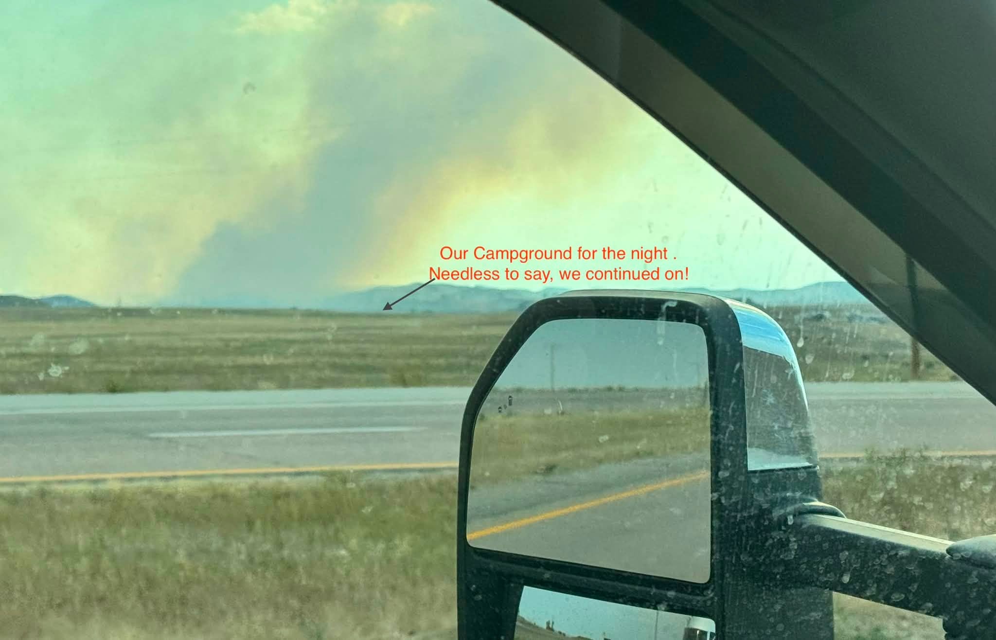 A column of wildfire smoke rising over the Wyoming prairie, seen through the truck windshield past the side mirror