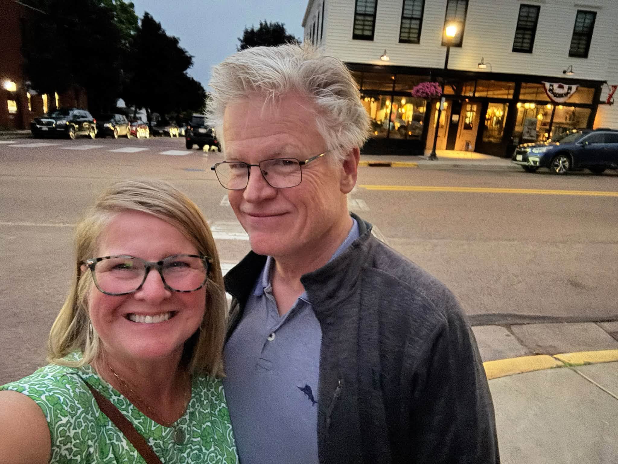 Sandra and Tom smiling on a Bayfield street corner at dusk, with lit storefront windows and a bunting-draped building behind them
