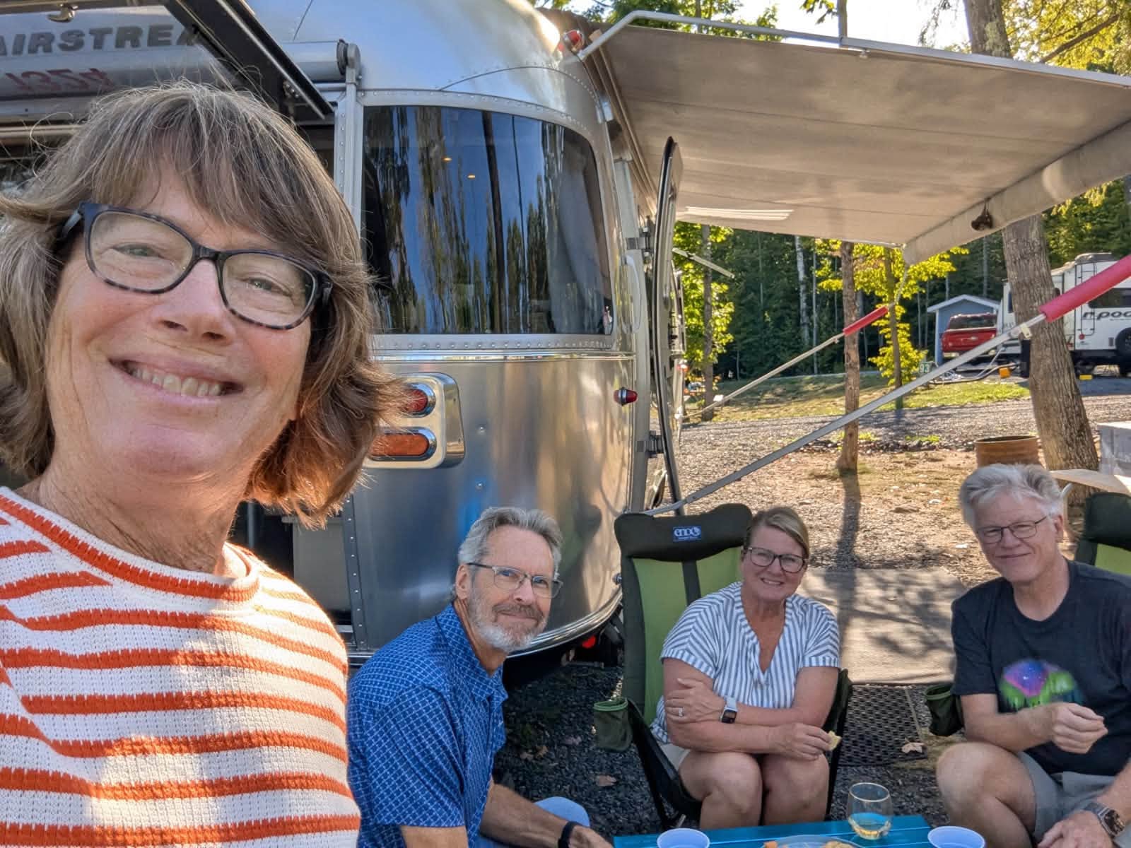 Four friends in camp chairs under Belle’s awning in the late afternoon sun, the polished Airstream filling the frame behind them