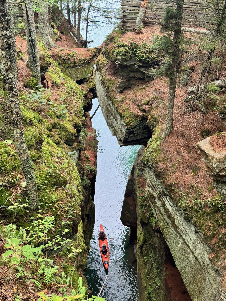 An orange kayak seen from directly above, threading a narrow slot of water between two moss-covered red rock walls