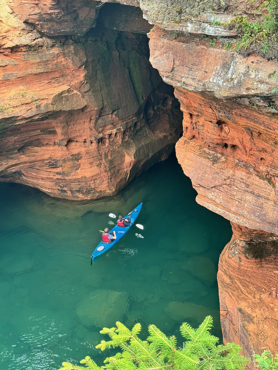 A blue tandem kayak floating in turquoise water at the mouth of a large sea cave carved into layered red sandstone