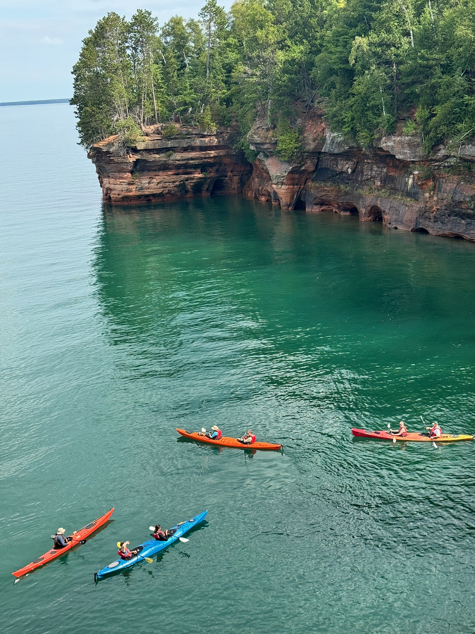 Looking down from the clifftop trail at four kayaks crossing clear green water below a pine-topped headland of red sandstone